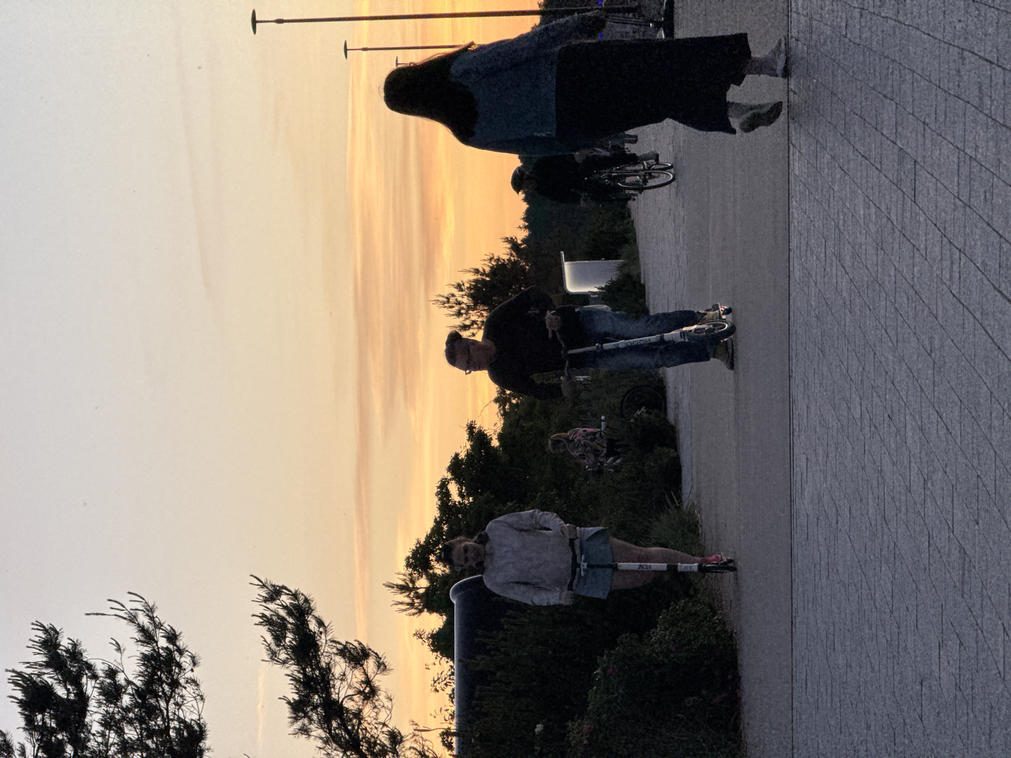 A group of people are enjoying an evening stroll or ride along a paved pathway in what appears to be a coastal or park area with lush green vegetation bordering the path. The sky is painted with the warm hues of a sunset, with soft oranges and yellows blending into a pale blue. In the foreground, a woman on a scooter is riding towards the viewer, dressed in a light-colored hoodie and a denim skirt. To her right, a man on a scooter is moving away from the viewer, wearing a dark top and jeans. Further in the background, another person is cycling on a bicycle. The pathway is bordered by streetlights, suggesting an urban or developed recreational area. The overall atmosphere is serene and pleasant, with the natural beauty of the sunset complementing the leisurely activities of the people. The location is identified as the city of Tallinn, Estonia, likely a scenic spot popular for evening recreation.