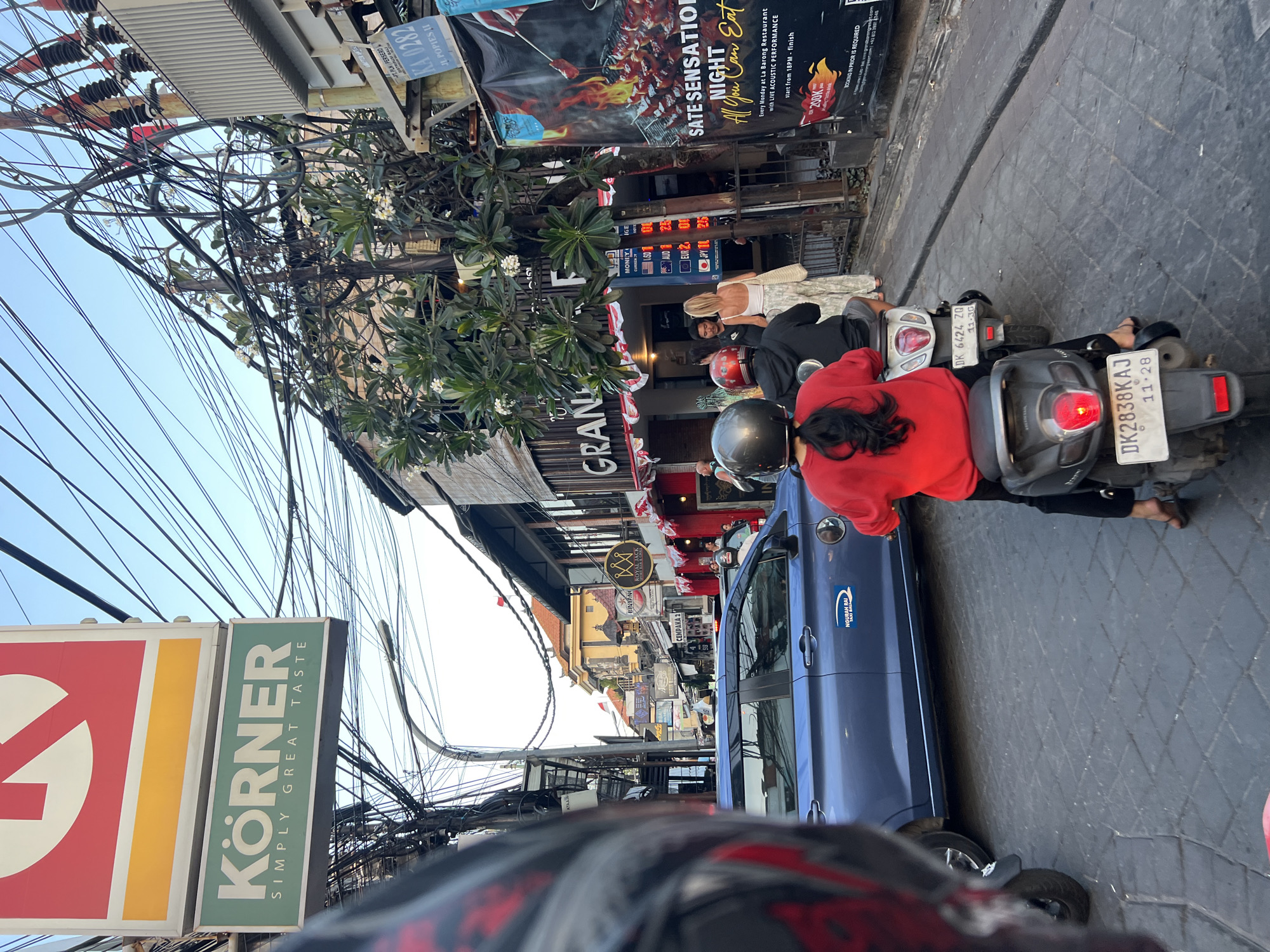 In Kuta, Indonesia, a street scene unfolds with several people on motorbikes, forming a slow-moving traffic line. The camera is positioned from the perspective of someone on a motorbike, partially obscuring their own helmet in the foreground. A blue car is stopped to the left, and a woman in a red top and black pants, riding a scooter, is a central figure. To her right, another person on a motorbike is also visible. In the background, commercial establishments line the street, with one featuring a large banner advertising "SATE SENSATION NIGHT All You Can Eat." Overhead, a dense network of electrical wires crisscrosses the sky, characteristic of many Southeast Asian cities. The road surface is paved with interlocking grey bricks.