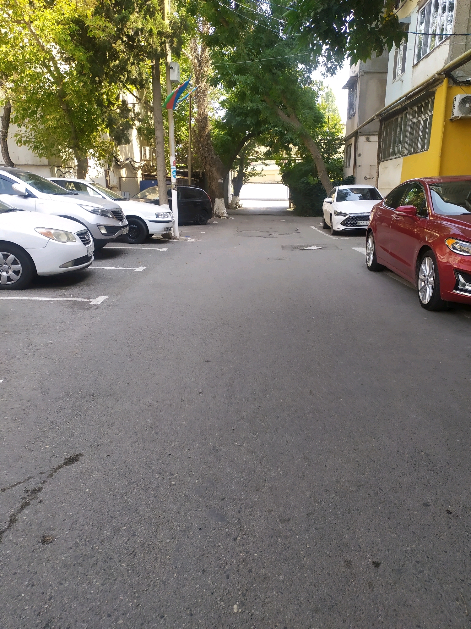 The image captures a street scene in Binagadi, Azerbaijan, with several cars parked on an asphalt surface. In the foreground, a row of white and silver cars is visible, parked in designated spots marked by white lines. Further down the street, a white car with a license plate starting with "AZ77" is parked, next to a dark-colored vehicle. On the right side of the street, a red car is parked, and behind it, a white car is also visible. The street is lined with trees, some of which arch over the road, creating a canopy. Buildings are visible on the right side of the street, with windows and an air conditioning unit. A flagpole with a flag, possibly the flag of Azerbaijan, is also present. The overall environment suggests a residential or commercial street in a city. There are no people visible in the image.