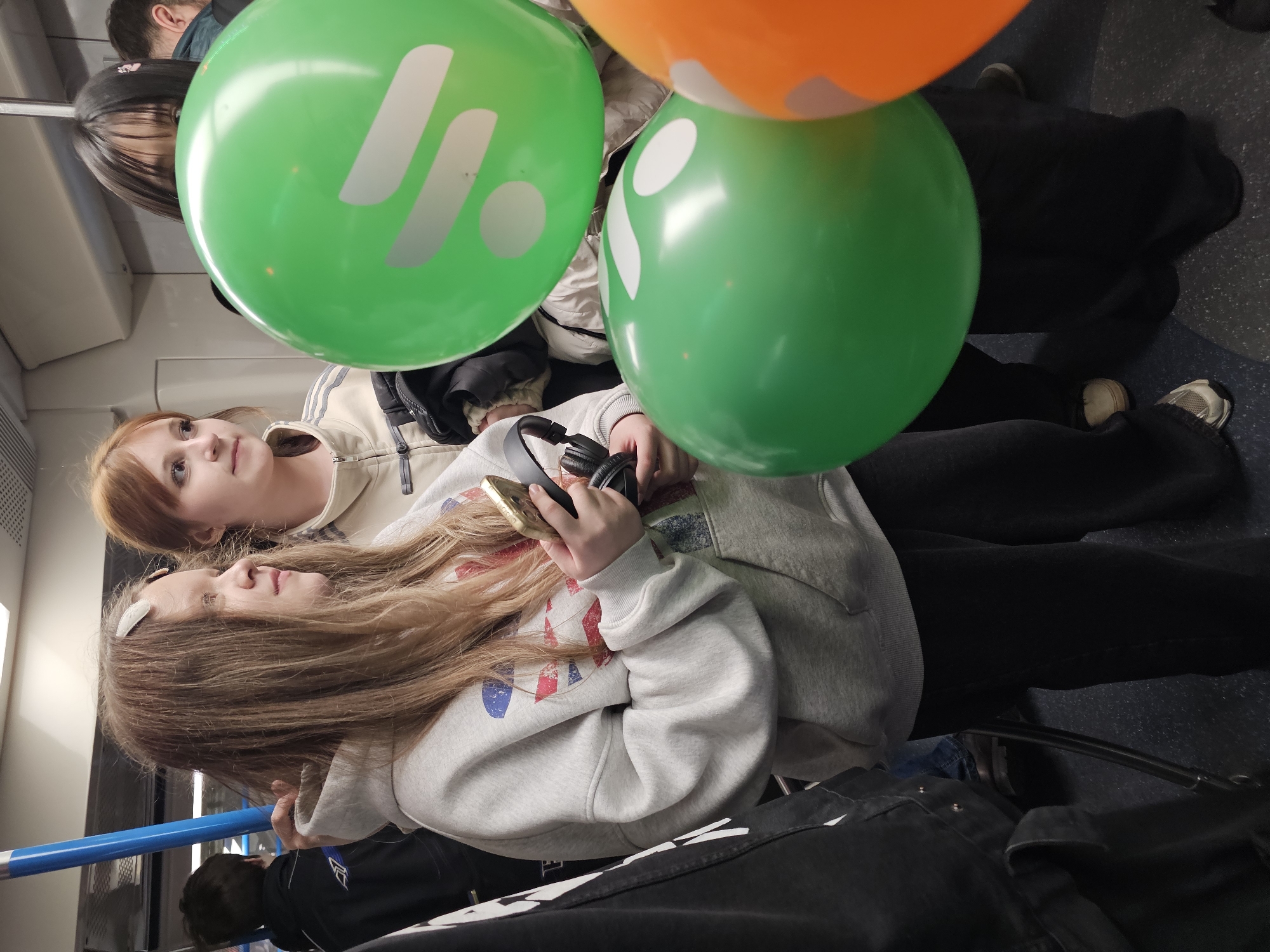 The image depicts a candid moment inside a public transport vehicle, likely a train or metro, with several passengers. The scene is rotated 90 degrees clockwise from its natural orientation.
In the foreground, two young women are prominent. The woman on the left (when viewed upright) has long, light brown hair and wears a light grey hoodie with a multi-colored graphic design, possibly resembling a flag. She is holding a gold-colored smartphone and a pair of black over-ear headphones. Her gaze is directed downwards or straight ahead. Adjacent to her, a woman with reddish-brown hair is dressed in a beige and white striped jacket, looking upwards and to the right with an observant expression.
Several balloons are central to the composition. Two prominent green balloons, each featuring a white, stylized logo composed of an angular shape (resembling an 'L' or '7') and a smaller circular or oval element, are positioned in front of and between the women. A vibrant orange balloon is also partially visible at the top of the frame.
Other passengers are vaguely discernible in the background, including a person with dark hair partially obscured by a green balloon, and another near a blue vertical support pole. The interior of the vehicle features light-colored walls and ceiling, and a dark, possibly patterned, floor. The presence of the balloons suggests the individuals might be traveling to or from a special event or celebration. The time of day is not evident from the indoor lighting.