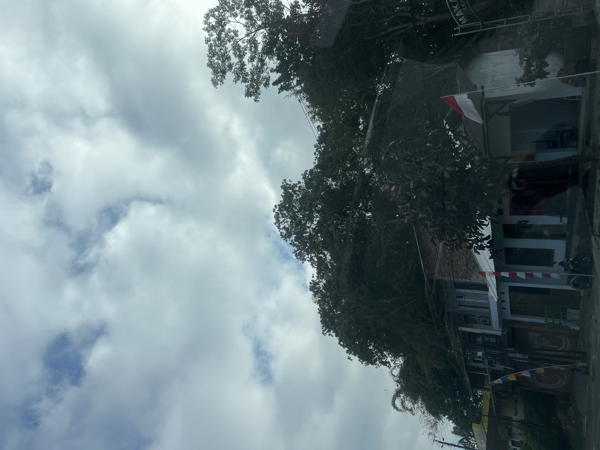 The image depicts a street scene in Kranggan, Indonesia, under a cloudy sky. In the foreground, a road is visible, with a motorcycle parked on the right side. A building with a white facade and a red and white flag is situated on the right. Adjacent to it is another building with a brick facade and multiple windows. Colorful flags, including one with blue, red, and white stripes, are displayed in front of this building. Dense foliage, including trees and bamboo, overhangs the buildings from a hillside in the background. A signpost with the word "MAKAN" is visible on the right side of the image. The overall atmosphere is overcast and appears to be daytime.