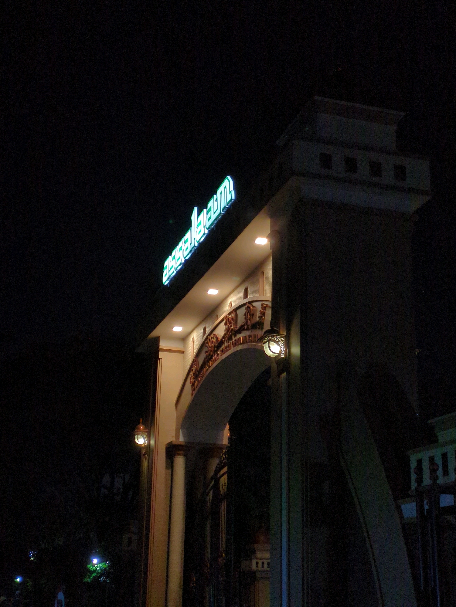 The image shows the entrance to a building at night, illuminated by neon lights and architectural lighting. A sign with the word "assalaamu" in green neon letters is visible at the top left of the frame. The entrance features an arched walkway with decorative relief carvings along the upper edge. Several light fixtures are mounted on the walls and pillars. In the background, the dark night sky is visible, with some distant lights and foliage. The architecture suggests a formal or ceremonial building. There are no people clearly visible in the frame. The image is a photograph taken at night, likely in an urban or semi-urban setting in Pabelan, Indonesia, given the context.