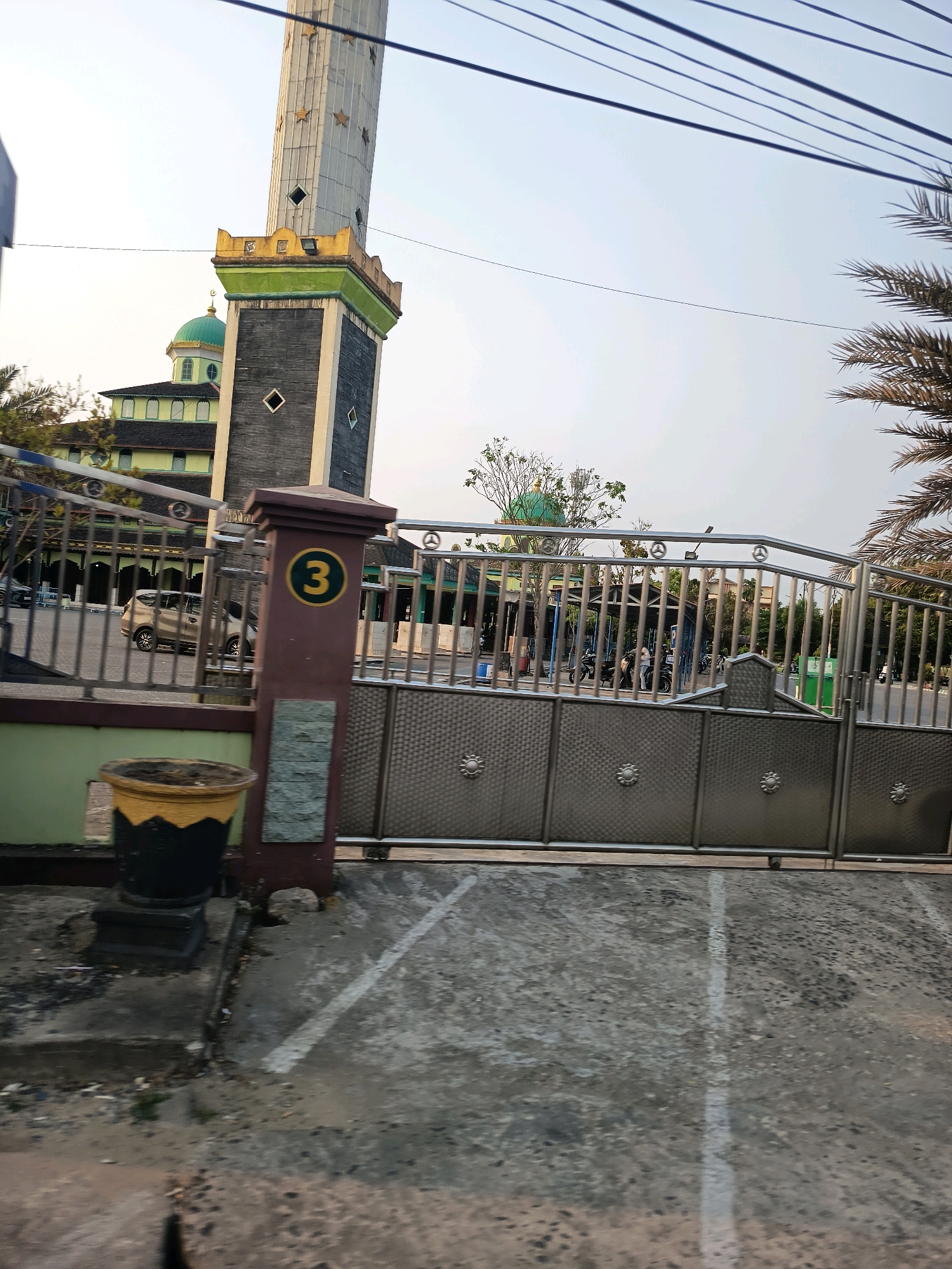 The image shows a mosque's minaret and dome, with a large, ornate metal gate in the foreground. A large concrete planter with a yellow and black rim sits to the left of the gate. A dark brown pillar with a yellow circle containing the number "3" is mounted on the left side of the gate. The gate itself is made of shiny metal with intricate patterns and decorative elements. Behind the gate, several motorcycles and a car are visible in what appears to be a parking area. A person is standing near the motorcycles. Palm trees and other foliage are present in the background. The sky is bright and overcast. This scene is likely located in the city of Banjarmasin, Indonesia, given the architectural style of the mosque and the presence of palm trees.