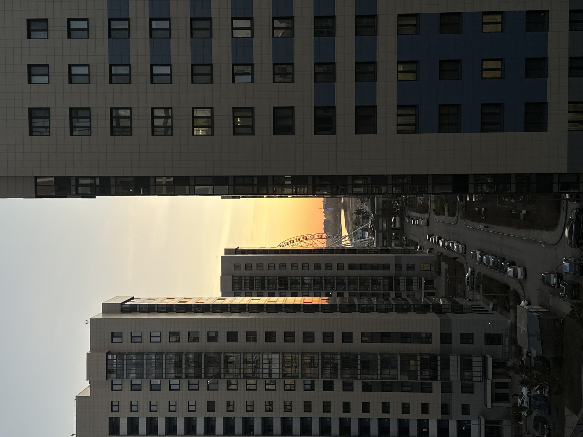 The image presents an urban landscape, likely captured during sunrise or sunset, as indicated by the vibrant orange and yellow glow in the sky. The photograph appears rotated 90 degrees clockwise from its natural orientation.
Dominating the upper and lower sections of the frame are two modern, high-rise buildings. Both feature grid-patterned facades constructed from light grey tiles, punctuated by numerous dark, rectangular windows. The building in the upper section additionally displays distinctive vertical stripes of blue cladding.
A wide horizontal band of sky spans the middle of the image, separating the two main building structures. The sky transitions from a pale grey near the top building to a brilliant orange and yellow at the horizon, casting distant elements into silhouette. Against this brightly lit sky, a large Ferris wheel is prominently visible, its structure forming a dark outline. Below the horizon, a body of water is partially discernible, followed by a street with numerous cars parked along its side. Further buildings and some sparse vegetation line the street.
There are no people or visible text in the image. The clear sky suggests fair weather. The presence of the Ferris wheel, especially in the context of Kazan, Russia, strongly suggests the landmark "Wheel of Review" at the Kazan Riviera.