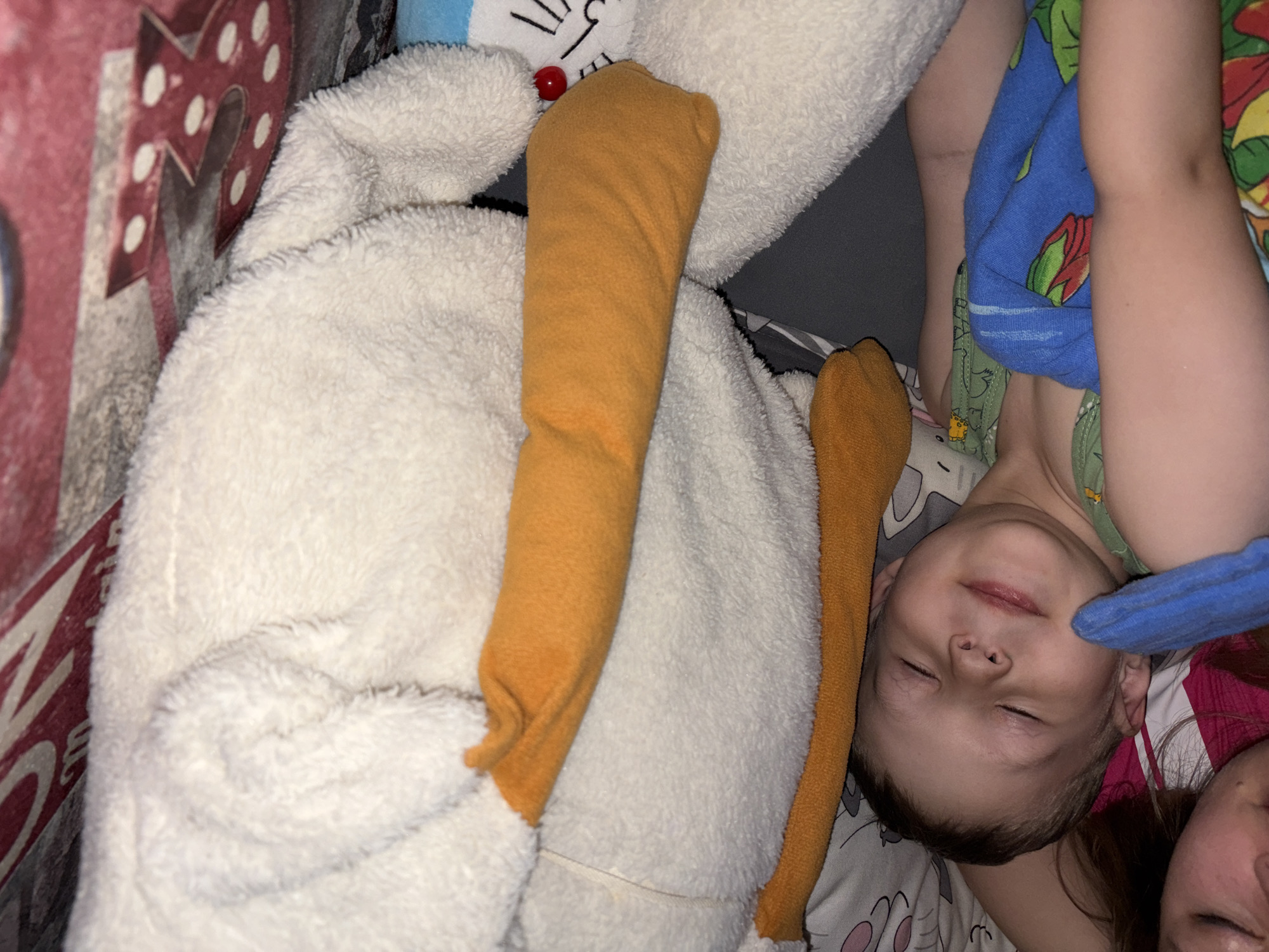 A child is asleep in a bed covered with a red and white patterned duvet. The child is wearing a green shirt and has a blue towel draped over them. Next to the child is a large white stuffed animal with orange appendages. Part of an adult’s face is visible in the lower right corner of the frame. The location is Chaikovsky, Russia.