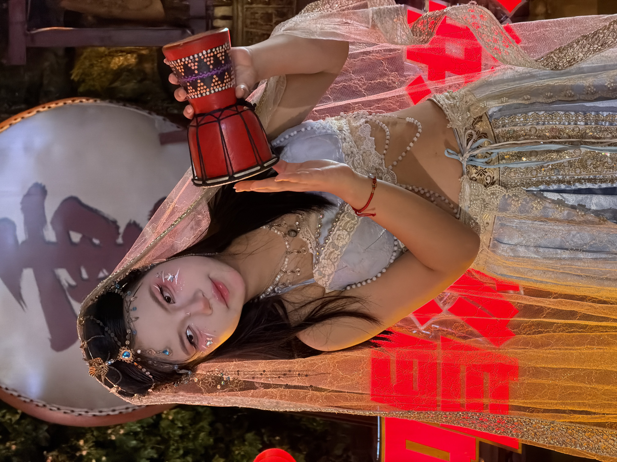 A young woman in traditional attire poses with a small drum. She is adorned with intricate jewelry, including a jeweled headband and a pearl necklace. Her face is decorated with subtle glitter accents. She wears a light blue and gold dress with ornate lace and sequined detailing. The scene is set against a backdrop of red Chinese characters, possibly part of a festival decoration, and a large drum with some indistinct imagery. The lighting suggests it is evening or nighttime. The woman's expression is serene and captivating.