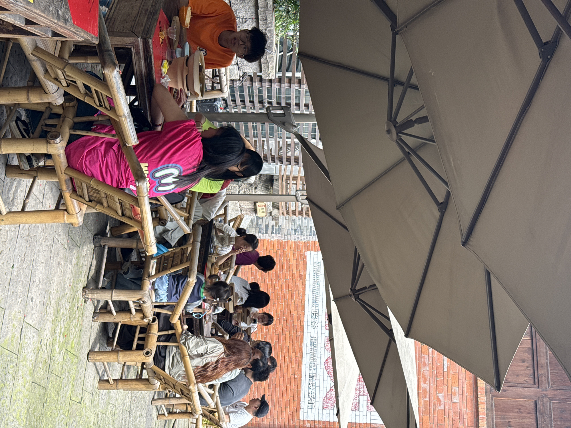 A group of about ten people is seated outdoors at tables with bamboo chairs, under the shade of large beige umbrellas. The people appear to be engaged in an activity, possibly a workshop, as some seem to be working with clay or other materials on the tables.
The location appears to be in the Yuzhong District of China, indicated by the Chinese characters written vertically on the brick wall behind them. The architecture includes brick buildings and the environment suggests a tourist or historical area. The lighting is bright, suggesting a sunny day.