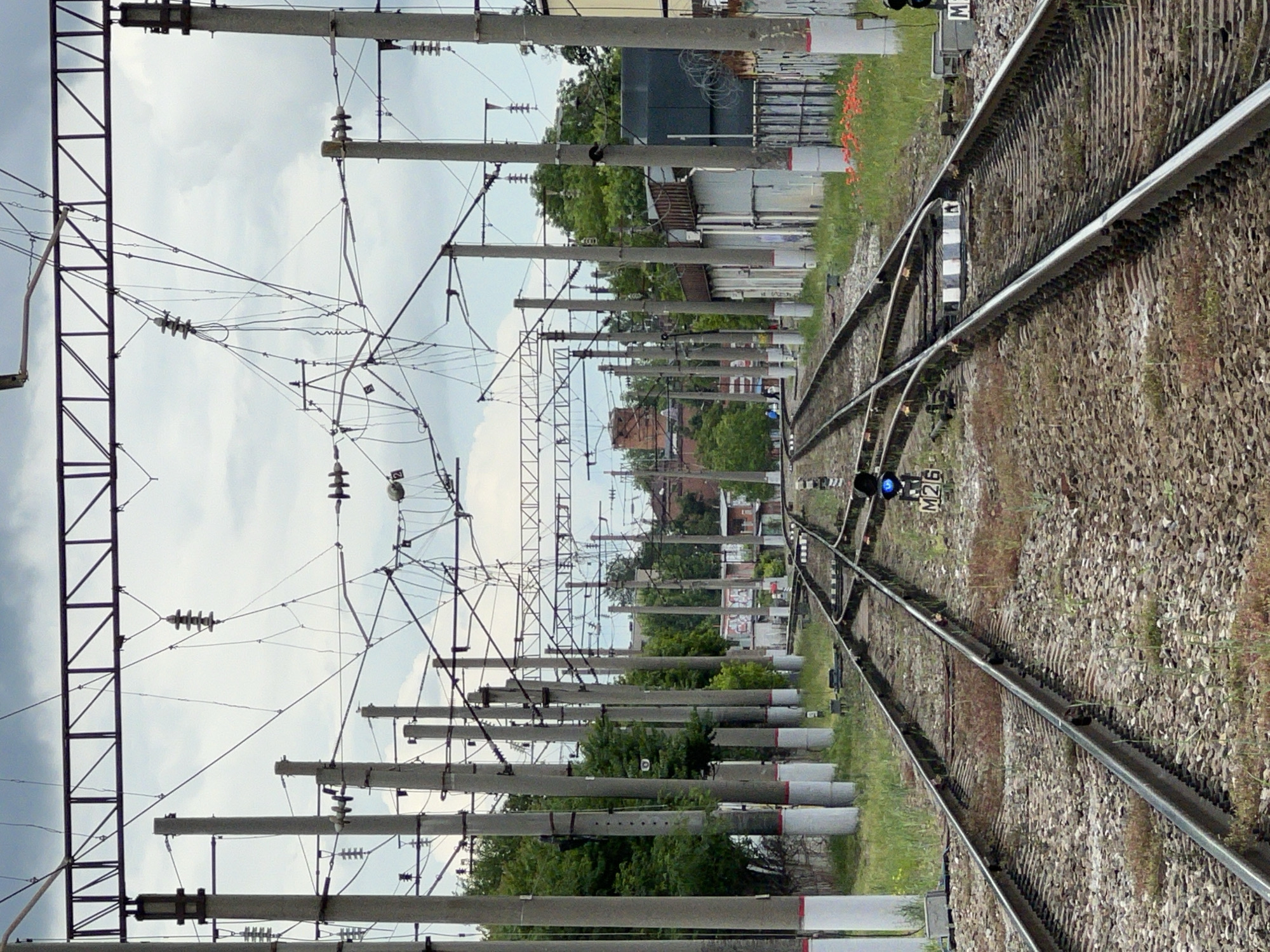 The image shows a railway track in Krasnodar, Russia. The tracks are surrounded by gravel and grass, with overhead power lines and poles running along the side. There is a blue signal light. Buildings and trees are visible in the background.