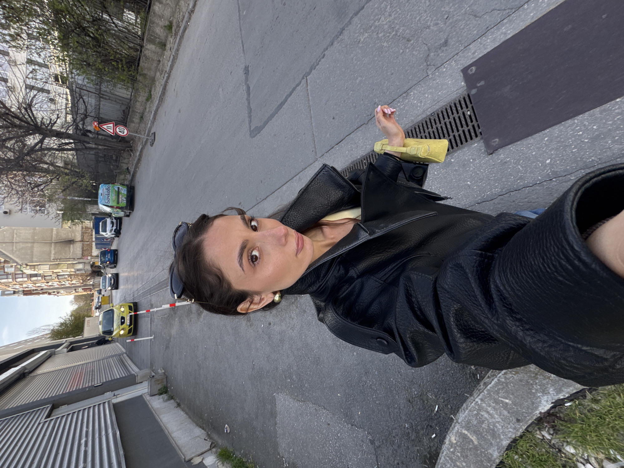 The image captures a young woman in an urban street setting, appearing to take a selfie. She is positioned on the right side of the frame, looking directly at the camera with a neutral expression. She has dark hair, dark eyes, and is wearing a black leather jacket over a lighter top, possibly yellow. Sunglasses are perched on her head, and she has gold-toned earrings. In her right hand, she holds a small, light yellow item, possibly a bag or phone case, and her fingernails are painted light pink.
The scene depicts a gray asphalt road running left from the woman, with a sidewalk and curb in the foreground. Vehicles are visible parked or driving down the street. Notable among them is a white van with the text "Бочково" (Bochkovo) clearly visible on its side, and a bright yellow car further along. Traffic signs are present, including a triangular warning sign depicting two cars and a circular speed limit sign indicating "40".
In the background, a mix of buildings can be seen, from older residential structures with numerous windows to a building with corrugated metal siding. Trees, some bare and others with fresh green leaves, line the street. The ground features a large drainage grate near the woman. The ambient light suggests it is daytime, possibly overcast. A number "3115" is faintly visible on a building wall in one of the crops. The presence of the "Бочково" brand text aligns with the provided context of Varna, Bulgaria.