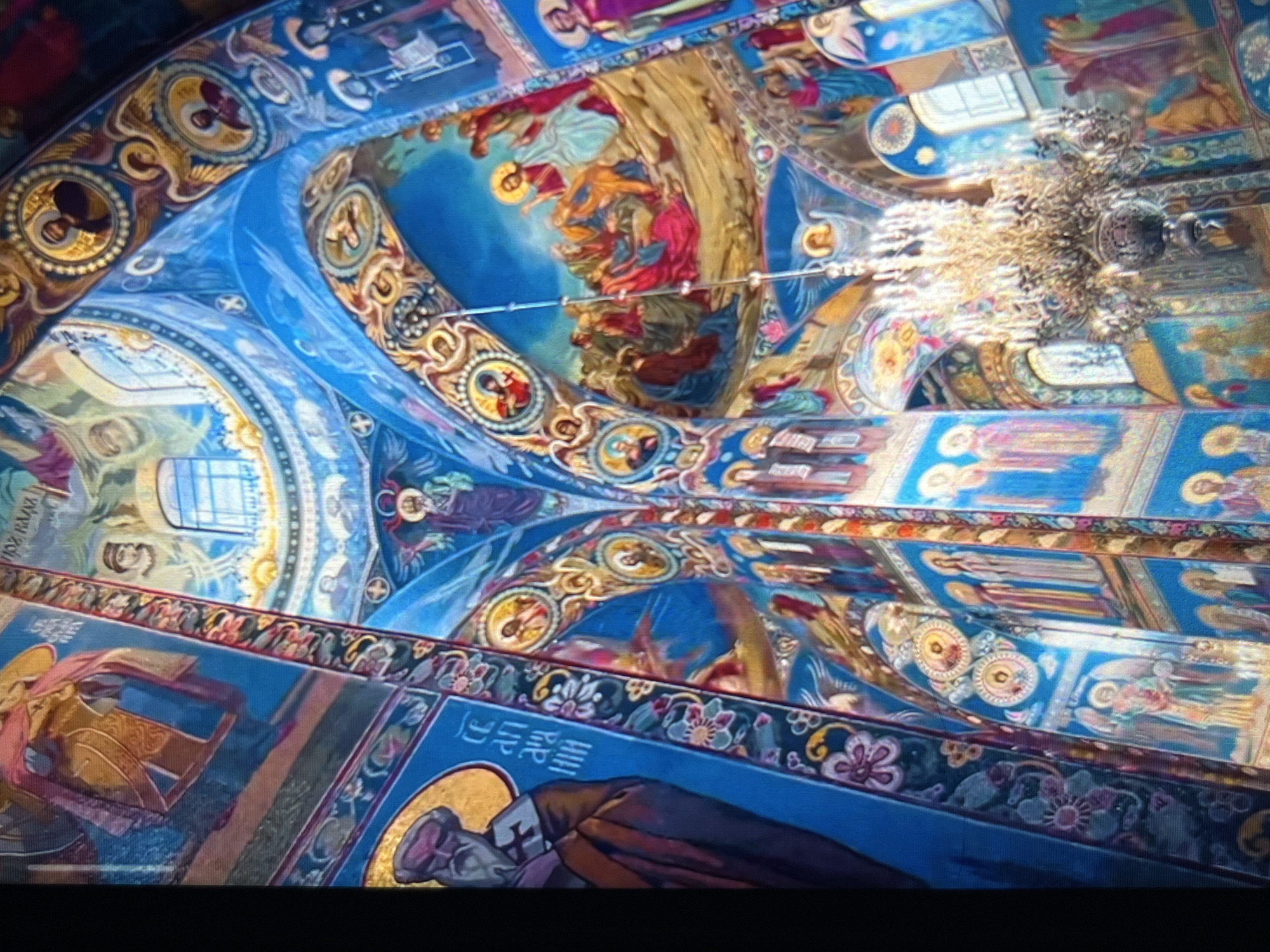 The image shows a highly ornate, painted ceiling, likely inside a church or cathedral, featuring religious iconography. The dominant colors are blue, gold, and red. Numerous figures and scenes depicting biblical stories are visible. A large chandelier hangs from the ceiling. The architectural style appears to be Eastern Orthodox or Byzantine. The ceiling exhibits decorative elements with circular patterns. The location is not Tinaquillo, Venezuela, but is instead Church of the Savior on Spilled Blood, St. Petersburg, Russia.