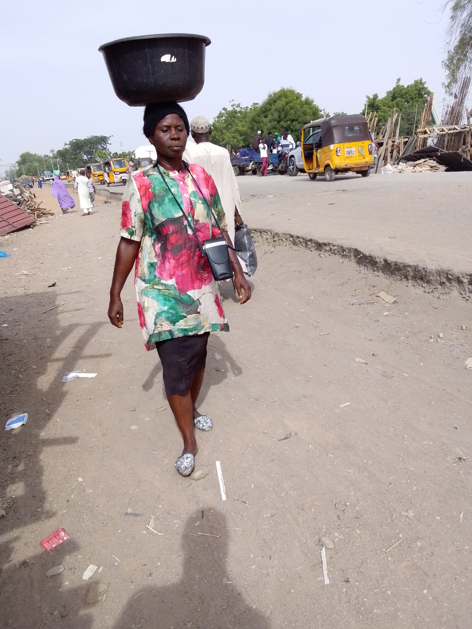 A woman walks along a dirt shoulder, carrying a large black tub on her head, in Jimeta, Nigeria. She is wearing a floral shirt, black skirt, and sparkly flats. A small black bag hangs across her chest. In the background are several auto rickshaws, various vehicles, and a group of people near what appears to be construction debris. The road surface is cracked and broken. Litter is scattered on the ground in the foreground. The sky is overcast.