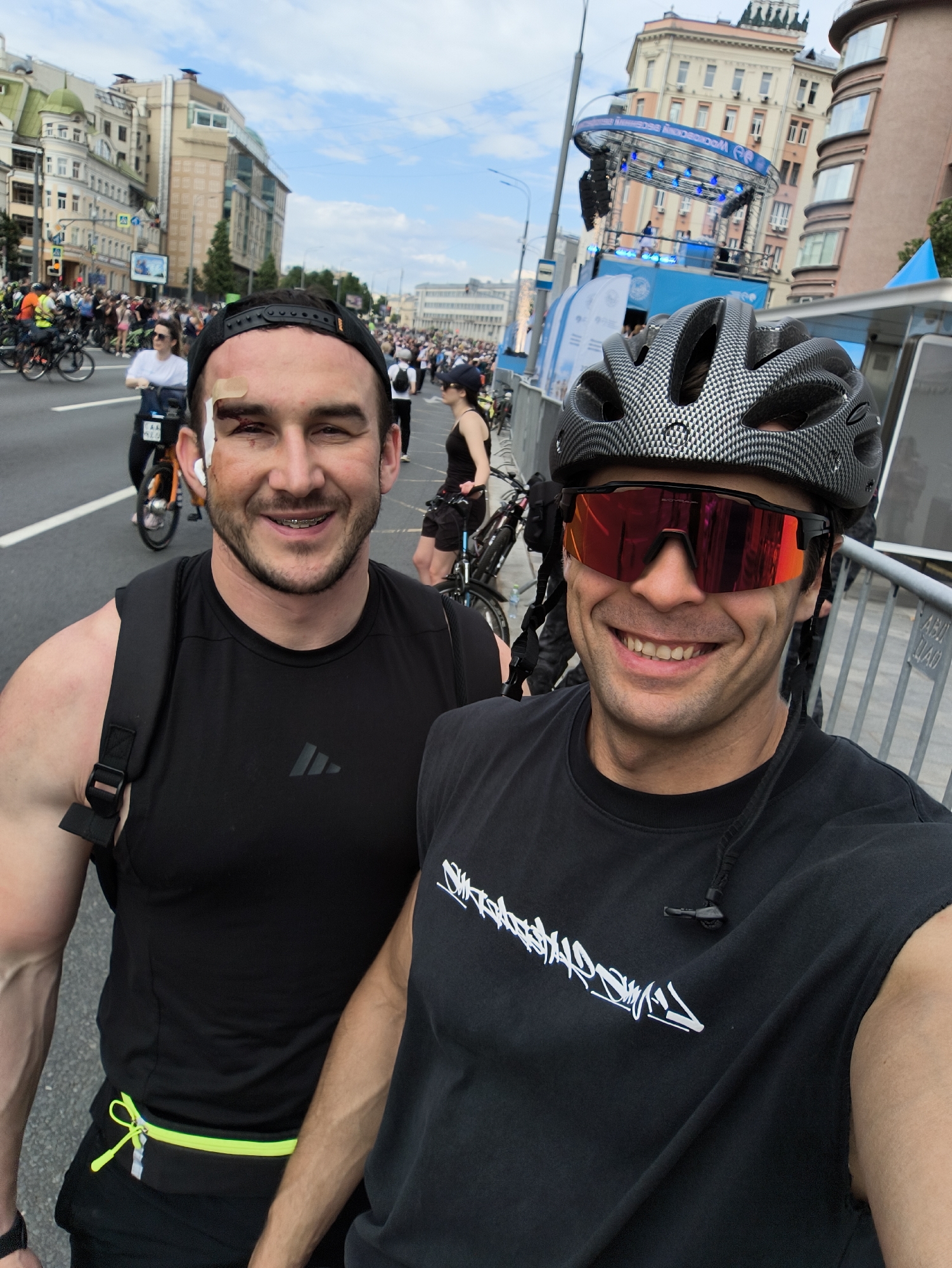 Two men pose for a selfie on a street in Moscow, Russia. The man on the left wears a black baseball cap, an Adidas tank top, braces, and a neon green running belt. He has a bandage on his forehead and bruises around his left eye. The man on the right wears a carbon fiber cycling helmet and red-tinted sunglasses. He wears a black t-shirt with a white graphic design. A crowd of people on bicycles fills the street behind them. A stage is set up in the background. Tall buildings line the street.