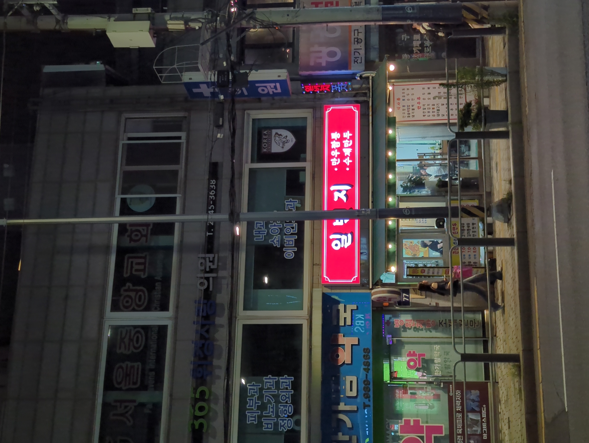 A nighttime exterior shot captures a city street scene in an urban environment. A tall, weathered utility pole stands prominently in the foreground, adorned with various wires and boxes, including a white, rectangular box with a red light on top. To the left of the pole, a building facade displays multiple illuminated signs and windows. A large, white sign with Korean characters "서울중앙교회" (Seoul Joongang Church) is visible near the top. Below this, a green sign reads "365 위생 사랑 의원" (365 Hygiene Love Clinic) with a phone number T. 245-3638. Further down, blue text on a white background indicates medical specialties such as "피부과" (Dermatology), "비뇨기과" (Urology), and "정형외과" (Orthopedics). A red, rectangular sign with white Korean text "일" (One), followed by "만두" (Dumpling) and "수제만두" (Handmade Dumpling), is also illuminated. To the right of the utility pole, another building features a blue sign with a white cross and Korean text "의원" (Clinic), and a purple sign with white text. A walkway with a black metal railing runs along the street, with a woman in dark clothing walking from left to right in the middle ground. She is wearing a black off-the-shoulder top and dark pants, carrying a pink shopping bag. In the background, a storefront with glass windows reveals people inside and illuminated menu boards. Potted plants are placed near the storefront. The street is paved with asphalt, and a sidewalk is visible.