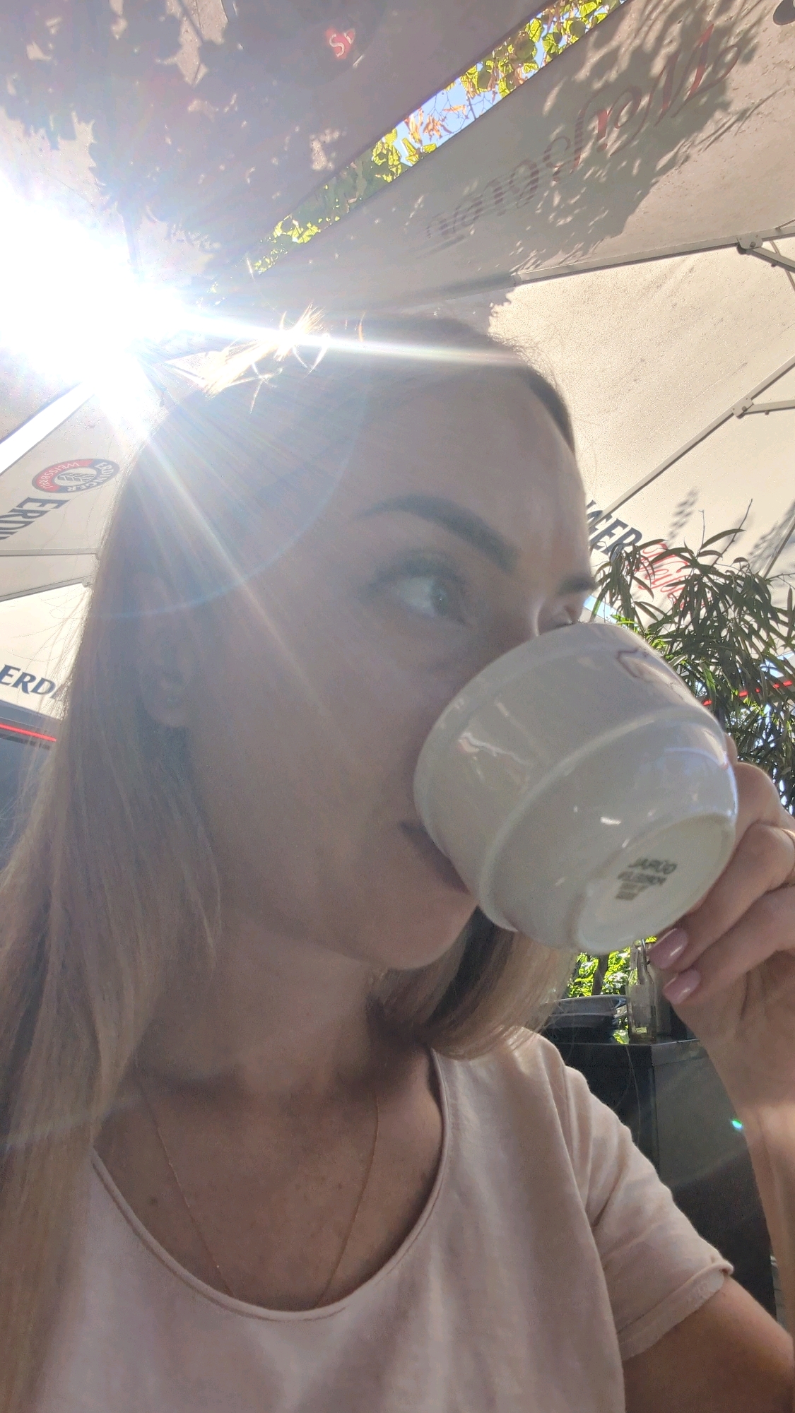 A woman with blonde hair and fair skin is captured in a close-up, profile view, drinking from a white ceramic cup. She is outdoors, possibly at a cafe, indicated by the presence of a large umbrella overhead, casting shadows of leaves and branches. Sunlight flares brightly into the lens from the upper left, obscuring details in that area. The woman is wearing a light-colored, V-neck top and a delicate necklace. Her fingernails are painted a light pink. In the background, a potted plant with green leaves is visible, along with what appears to be signage with text, partially obscured by the umbrella and sunlight. The overall scene suggests a casual outdoor setting. The context provided about Chișinău, Moldova, is not directly observable within the image itself.