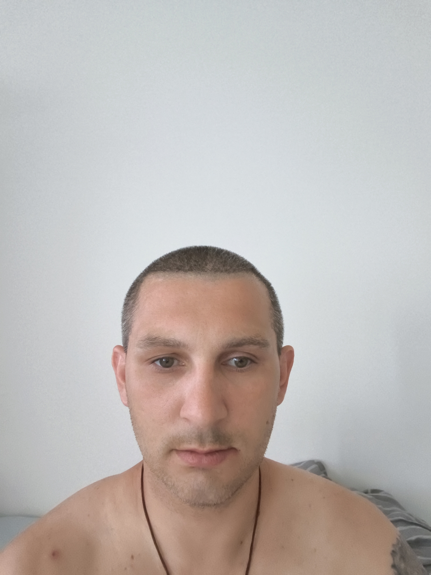 A man with a buzz cut and light stubble is pictured in a close-up, head-and-shoulders shot. He is facing the camera directly, his expression neutral. His eyes are open and looking forward, and his mouth is closed. He is shirtless, revealing his shoulders and a portion of his chest. A thin, dark cord is visible around his neck. The background is a plain, off-white wall. The image is lit evenly, with no strong shadows. The location is specified as Wolgast, Germany.