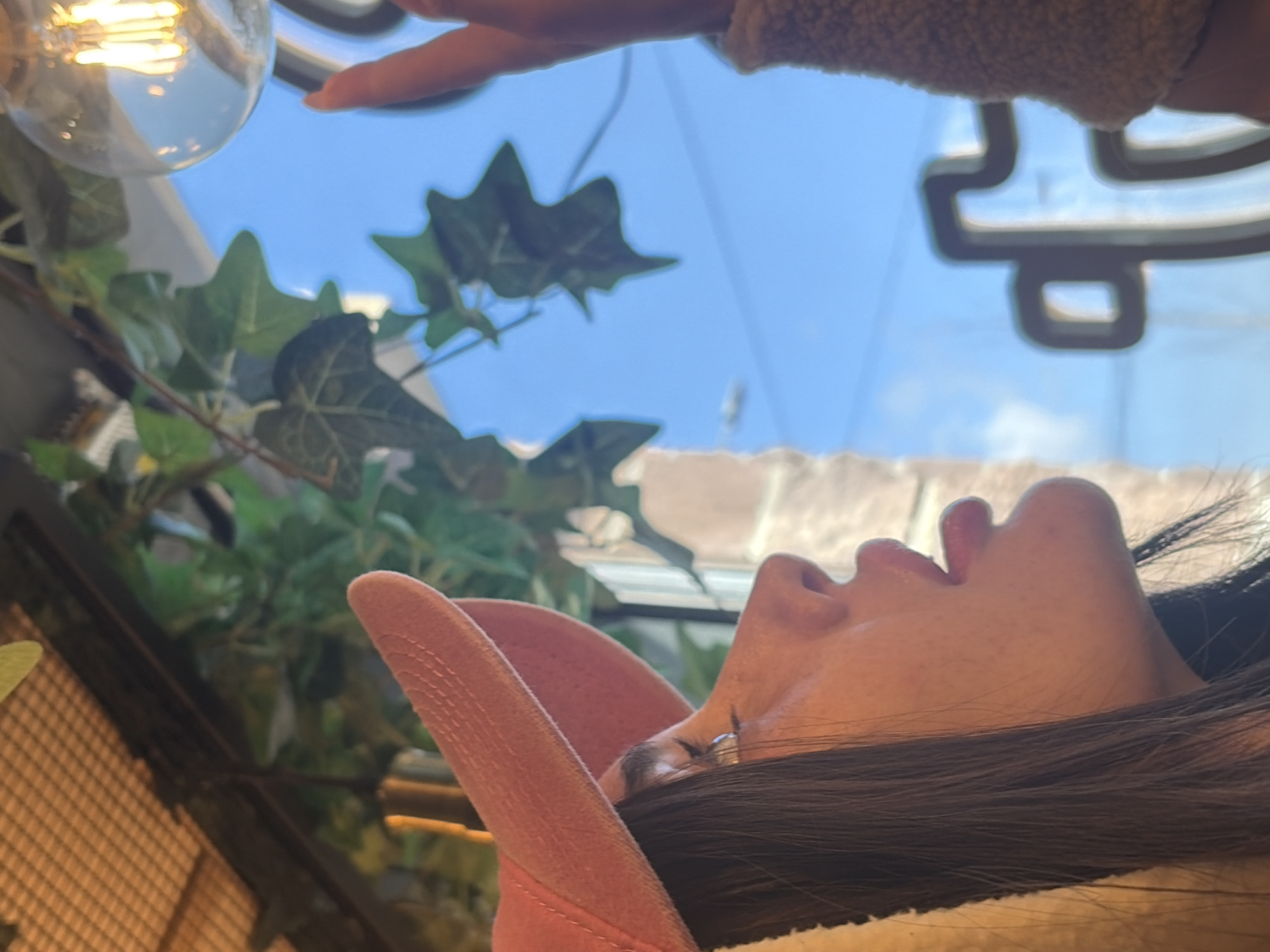 The image depicts a female individual in a café interior, seen in profile from the chest up. She wears a pink baseball cap with a finely ribbed texture and has long, dark hair. Her gaze is directed upwards towards a lit Edison-style light bulb. Her right index finger is extended and appears to make contact with the bulb. Her right arm is clothed in a light brown, textured garment. In the background, a large window reveals a clear blue sky, with a dark, stylized coffee cup logo, consistent with the Coffee Like brand, visible through the glass. Faux ivy plants are draped near the window, and a grid-like ceiling structure is present in the upper left corner. A reflection of a window frame is visible in her eye. The scene is located in a Coffee Like café in Saint Petersburg, Russia.