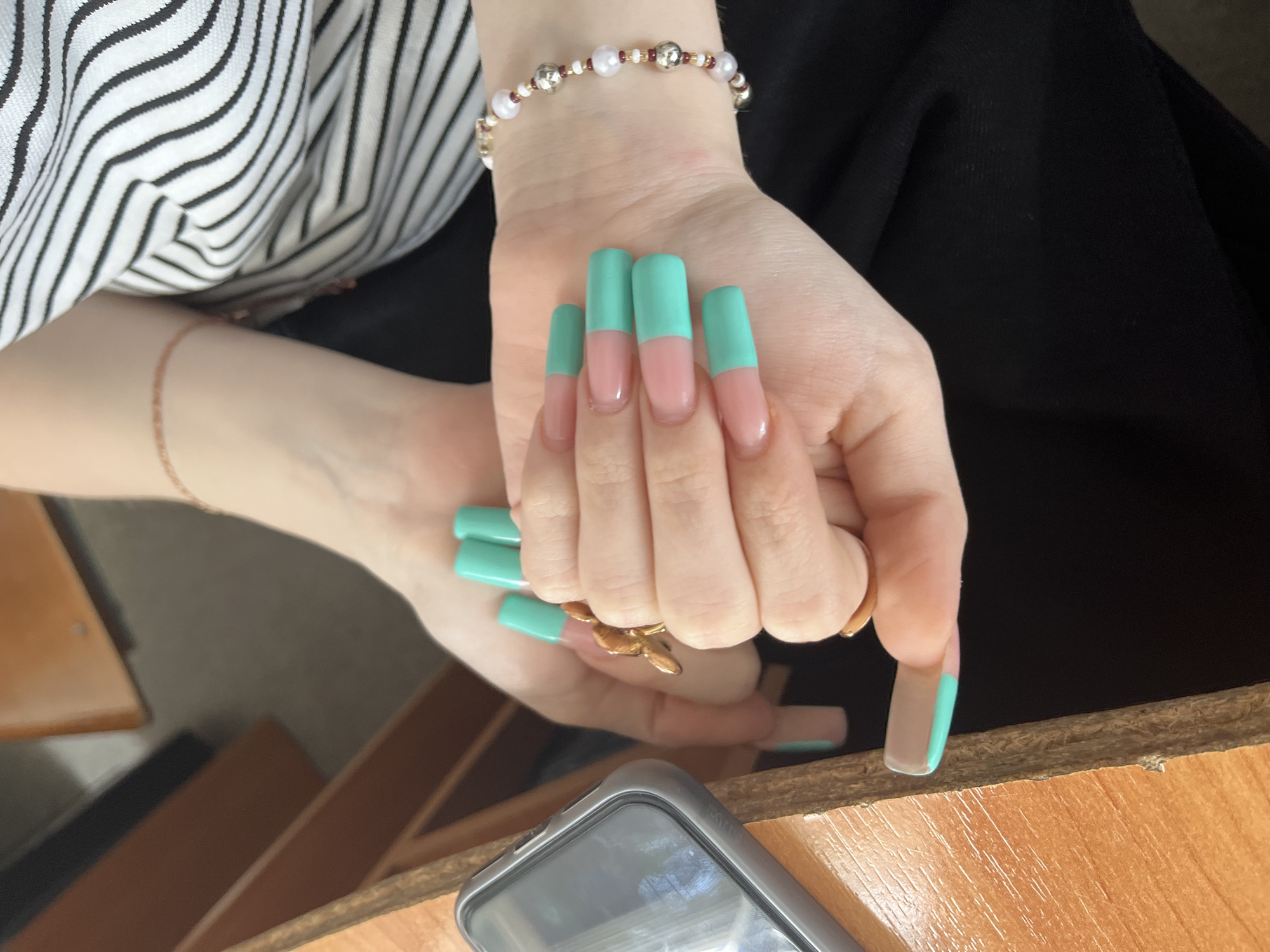 The image shows a close-up of a person's hands. The hands have long, square-shaped fingernails painted in a French manicure style with a sheer pink base and a bright teal tip. The person is wearing rings on two fingers and a bracelet on their wrist. The background is blurred, showing a wooden surface and a black garment. The context of Sterlitamak, Russia is not visually evident in the provided image.