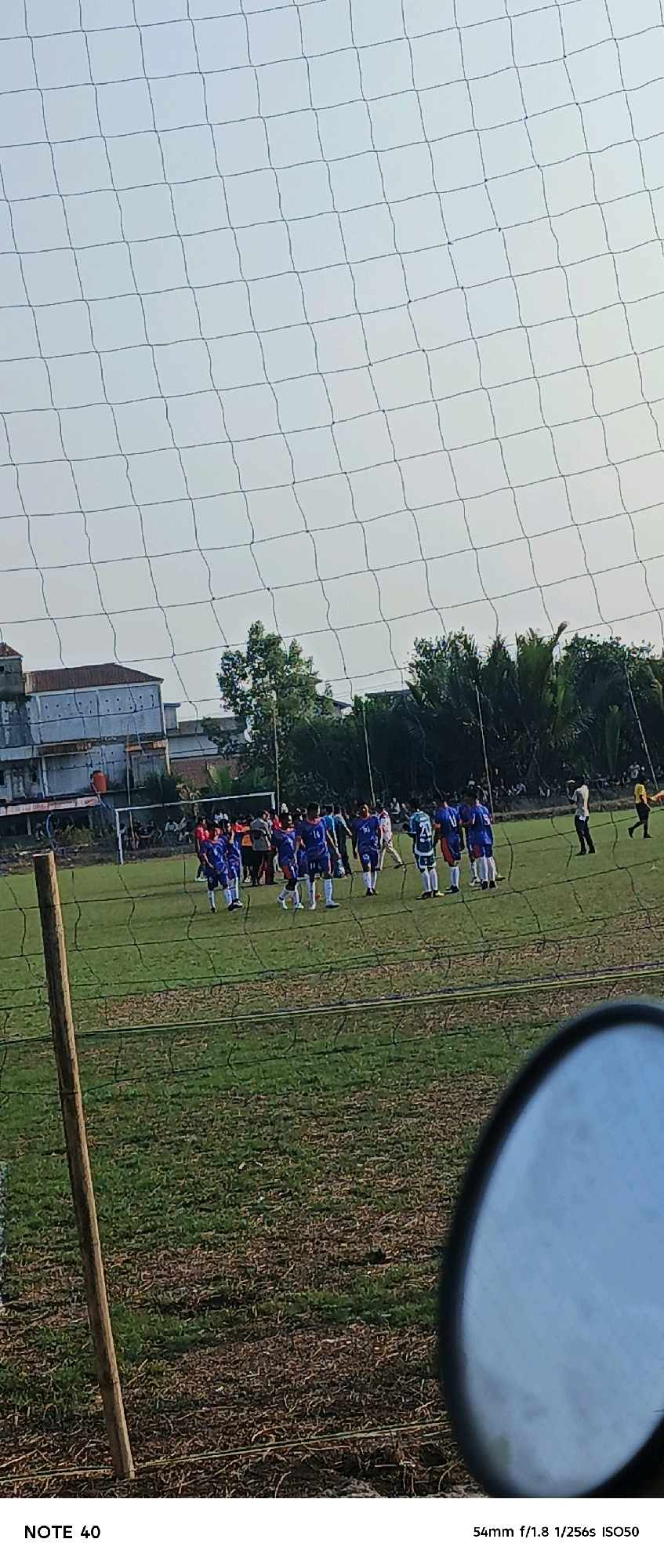 In Simbur Naik, Indonesia, a soccer match is in progress on a grassy field. A net is in the foreground, partially obscuring the view of the players. The players, some wearing blue uniforms, are scattered across the field, with a goal visible in the background. A few spectators can be seen in the distance. The sky is overcast, and trees line the edge of the field. A wooden post stands on the left side of the image. A circular reflection, likely from a camera lens or mirror, is visible in the bottom right corner.