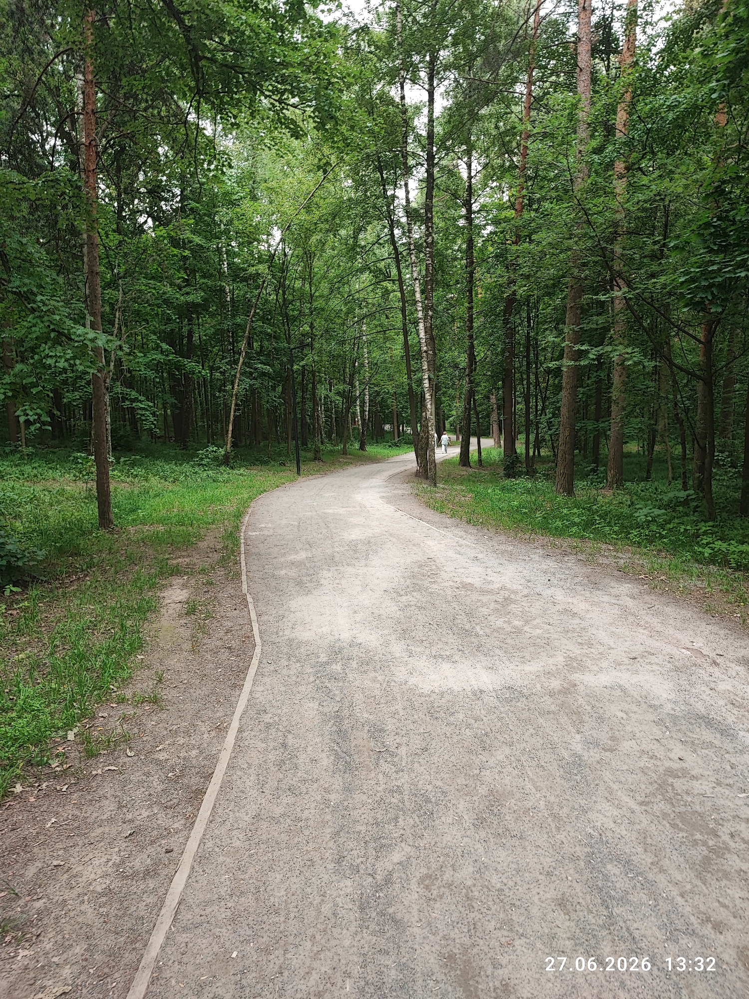 An eye-level, outdoor, vertical shot captures a gravel path winding through a dense forest. The path is on the right and curves to the left into the distance, bordered by a strip of concrete. Lush green grass and undergrowth flank the left side of the path. Tall trees with dense green foliage dominate the background, creating a shaded canopy. A lone figure in a white shirt and dark pants walks away from the camera on the path in the distance. The scene is dated 27.06.2026 at 13:32.