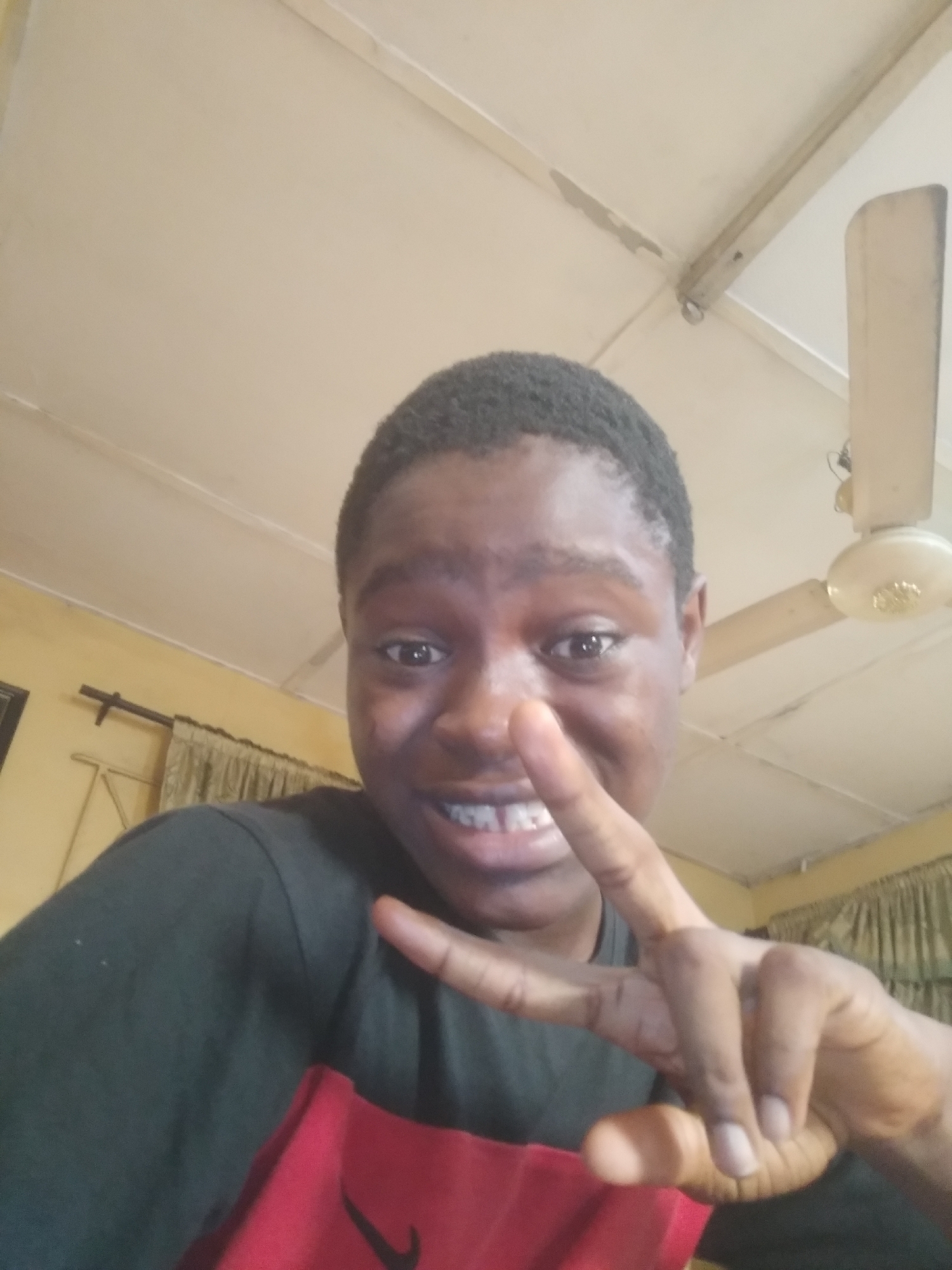 A young, dark-skinned person is holding up a hand in a "peace" or "victory" sign. The person has short, dark hair and is smiling, showing their teeth. They are wearing a dark green shirt with a visible red section featuring a Nike logo on the lower portion of the torso. The background includes a cream-colored ceiling with visible ceiling fixtures, including a ceiling fan. Part of a window with curtains is also visible. The image was most likely taken inside a residential area, and the exact location of the city of Shangisha, Nigeria cannot be determined based on the image provided.