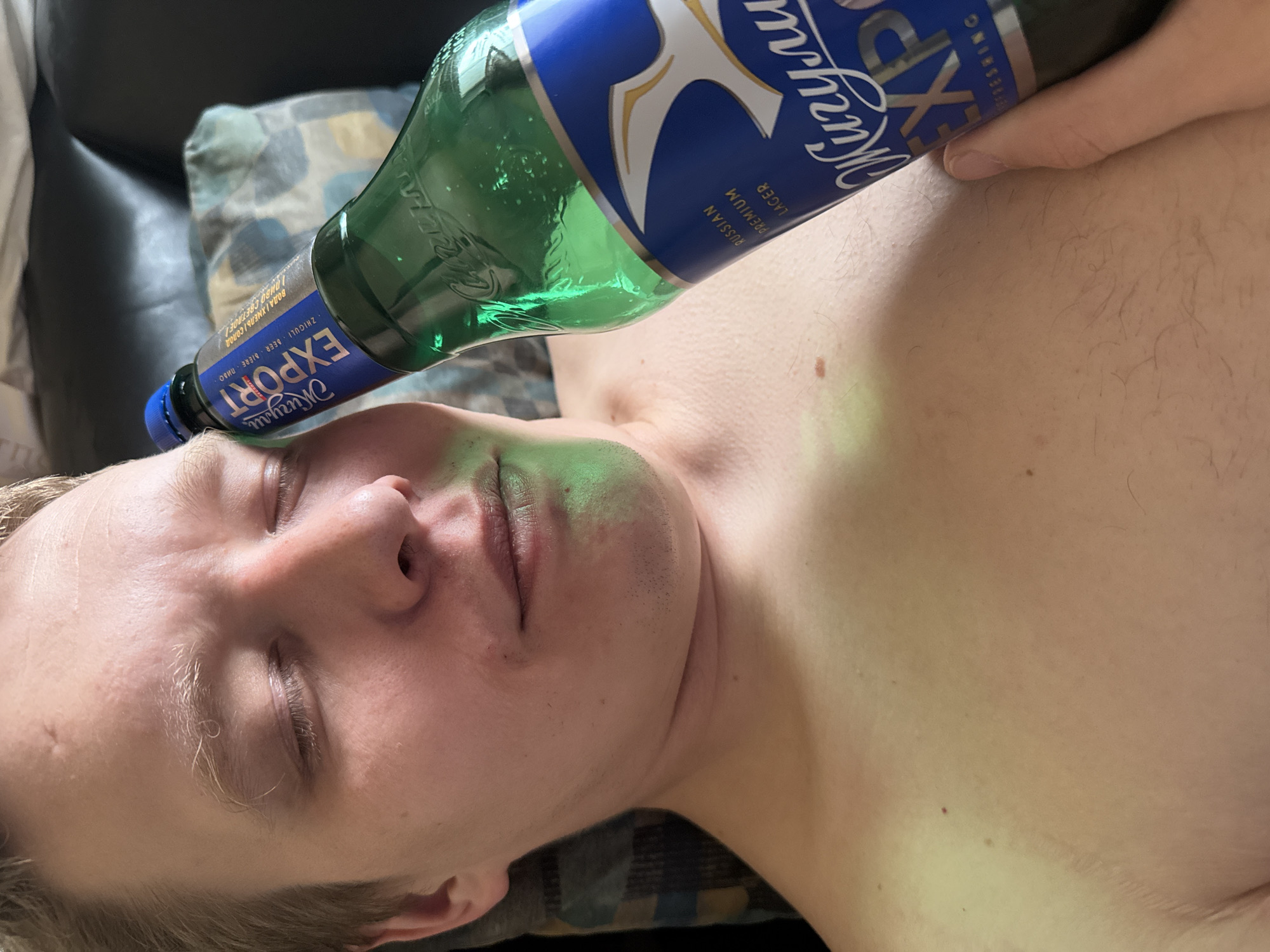 A man is lying down with his eyes closed, and a green beer bottle is being held over his face. The bottle is a "Zhiguli Export" Russian Premium Lager. The bottle has text on it in both Cyrillic and English. The beer's label indicates that it contains water, hops, and malt. The scene occurs on a patterned cushion or pillow. The location is not identified.