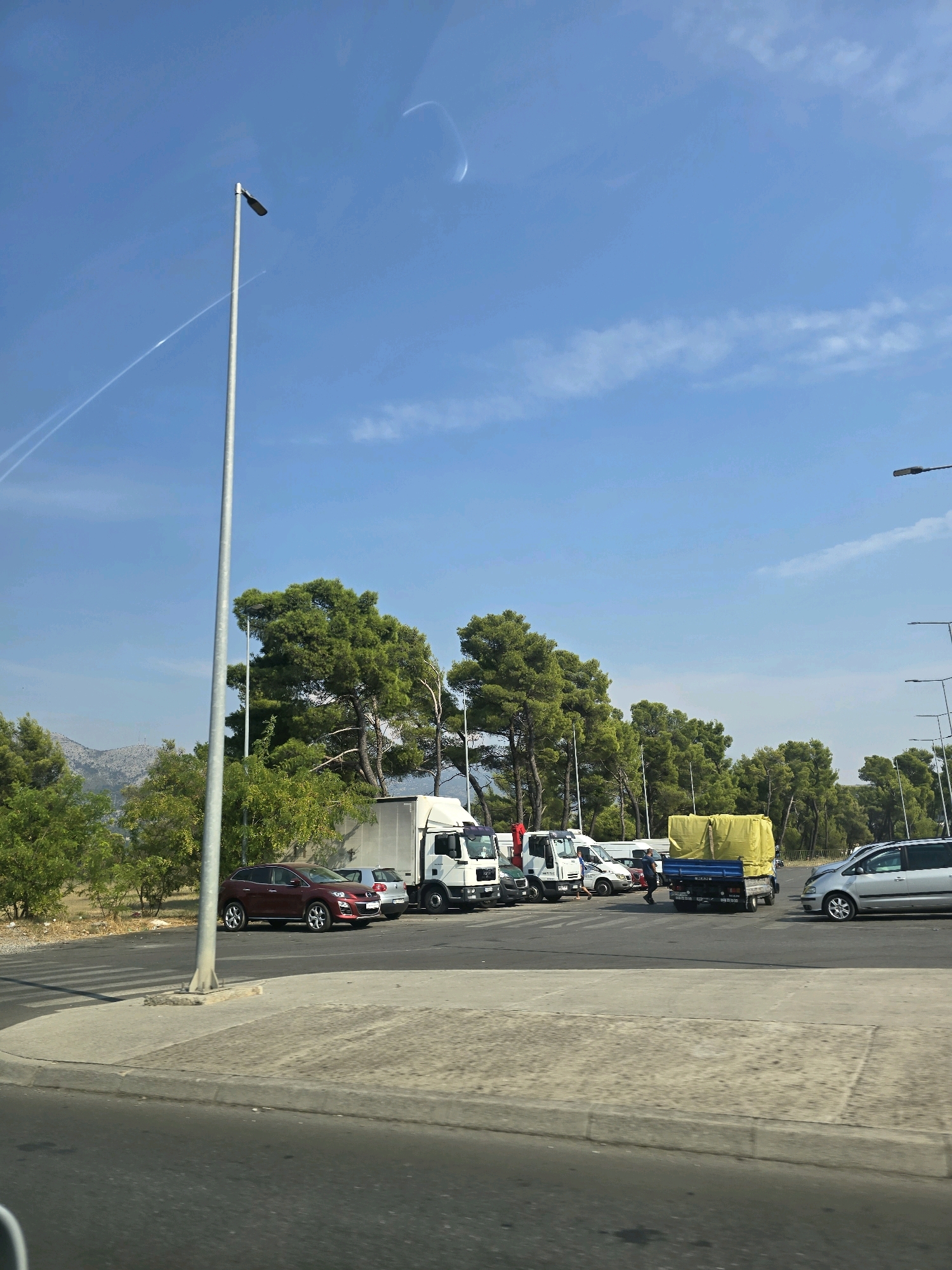 The image depicts a parking lot filled with various vehicles, including cars, trucks, and a van, set against a backdrop of lush green trees and a clear blue sky. A prominent lamppost stands on the left side of the frame. In the distance, mountains are visible under the bright sunlight. The scene appears to be a public area, possibly near a road or an industrial zone.