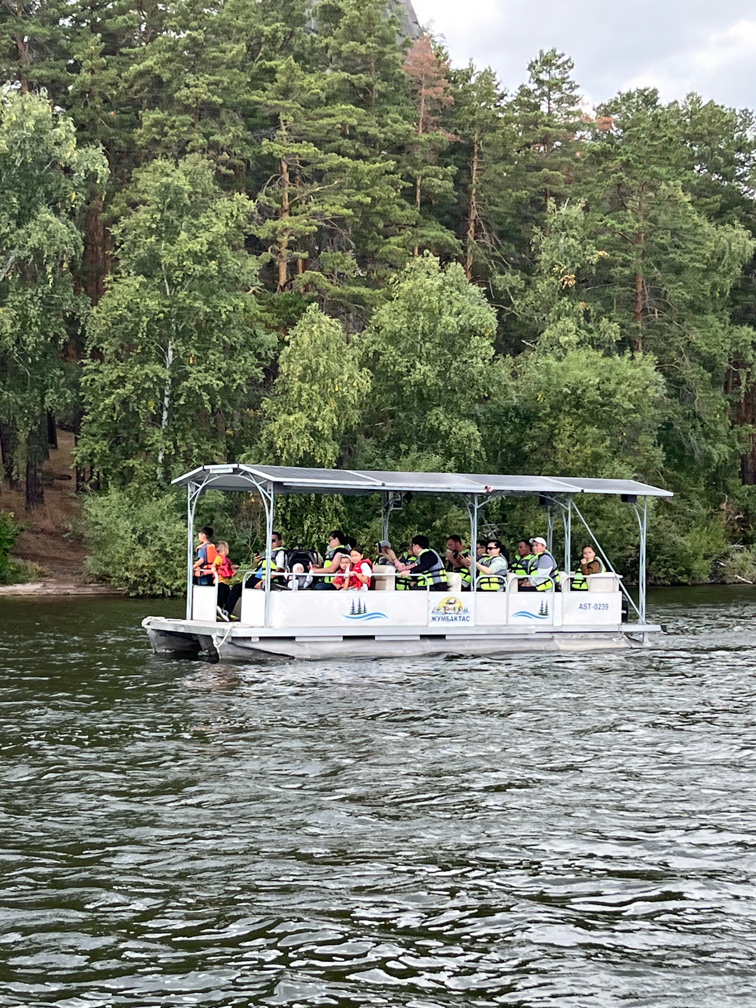 A pontoon boat with a canopy is carrying approximately ten people across a body of water. The boat has "ЖУМБАКТАС" and "AST-0239" written on its side. The people on the boat are mostly wearing life vests in yellow and orange. They are facing in various directions, some looking out at the water or the shore. The boat is moving from left to right in the frame. The background consists of a dense forest with tall trees and some undergrowth, suggesting a natural or park-like setting. The water is rippled, indicating a slight breeze or movement. This scene is likely at a recreational area within Зелёный Бор ауылдық округі, Kazakhstan, given the presence of the boat and the forested landscape.