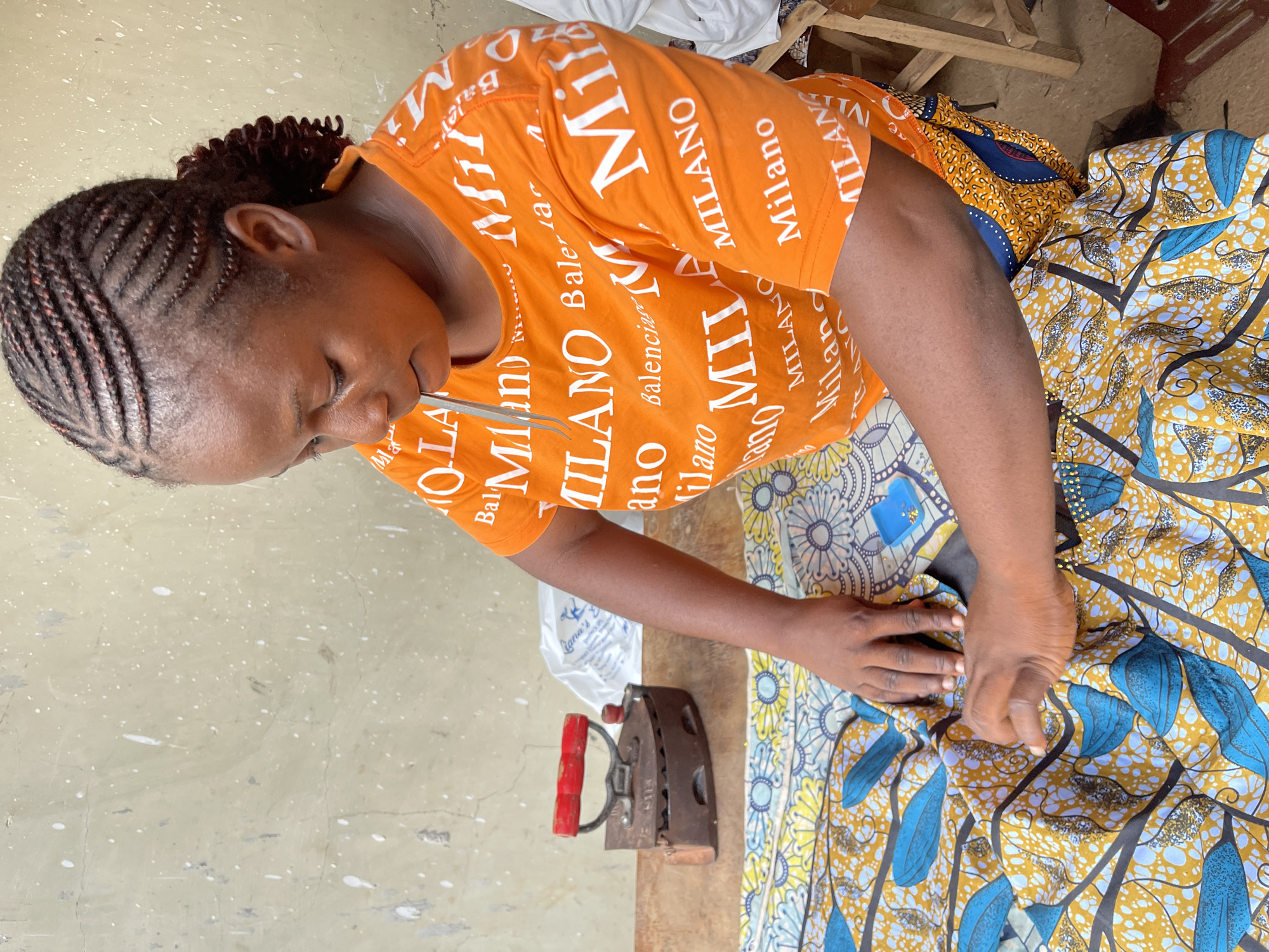 This media file shows a woman in Akwanga, Nigeria, working on a piece of fabric. She is wearing an orange t-shirt with repeated white text that reads "MILANO" and "Balenciaga," and a colorful wax-print fabric skirt with a yellow, blue, and black pattern. The woman has her hair braided and appears to be using tweezers to hold and manipulate the fabric. In the foreground, there is an old-fashioned, dark metal iron with a red wooden handle. The background is a textured, light-colored wall with white speckles, and a wooden surface with some bags or cloth visible. The lighting suggests it could be daytime. The overall scene appears to be an informal, perhaps domestic, workspace where the woman is engaged in sewing or tailoring.