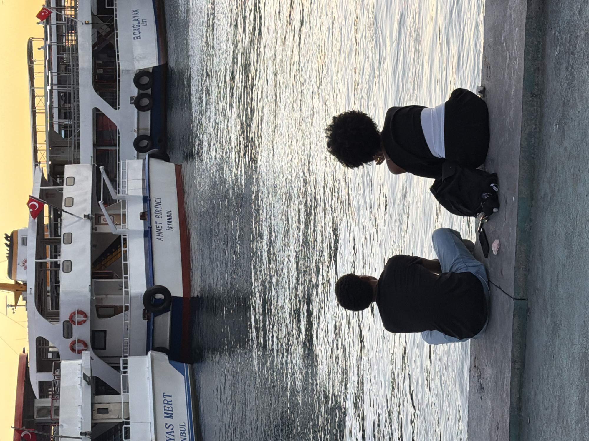 Two individuals sit facing away from the camera, looking out at the water. The person on the left has dark, short hair and wears a black t-shirt and light blue jeans. The person on the right has dark, curly hair and wears a black top with a white sash around their waist and black pants. They are positioned on a concrete ledge overlooking a body of water. Behind them, several ferries are docked. The ferries are white with red and blue accents. One ferry has the text "AHMET BİRİNCİ ISTANBUL" visible. Another has "B.CAGLAYAN LIST" visible. The sky above is a warm, orange-yellow, indicating either sunrise or sunset. A black backpack and a mobile phone are on the ledge near the individuals. The image appears to be taken in Kadıköy, Turkey, given the presence of ferries and the Turkish flags on the vessels, characteristic of Istanbul's ferry system.