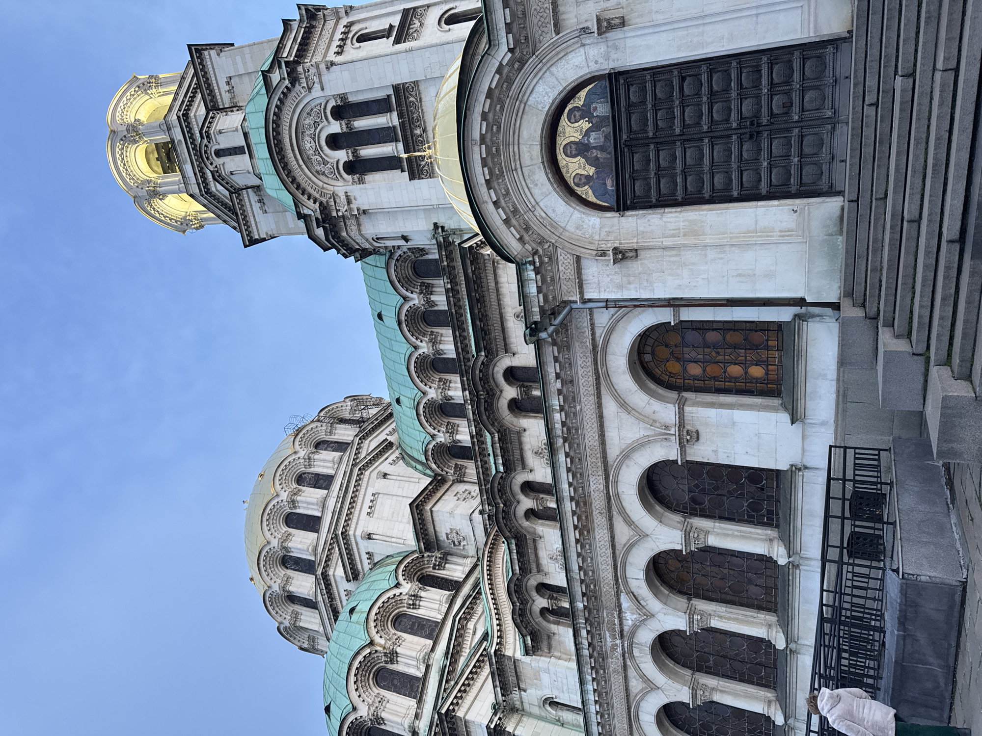 The image depicts the exterior of the Alexander Nevsky Cathedral in Sofia, Bulgaria. The view is angled upwards, capturing the intricate architectural details of the building. The cathedral is constructed of light-colored stone, with multiple domes topped with a green patina. Windows and arches punctuate the facade. The upper portions feature gilded domes and a bell tower illuminated by internal lighting. To the right, a large, dark door is set within an arched frame, above a series of arched windows. Steps lead upwards towards the door. A black metal railing and a figure wearing a white garment are visible in the foreground on the right. The background is a clear, blue sky.