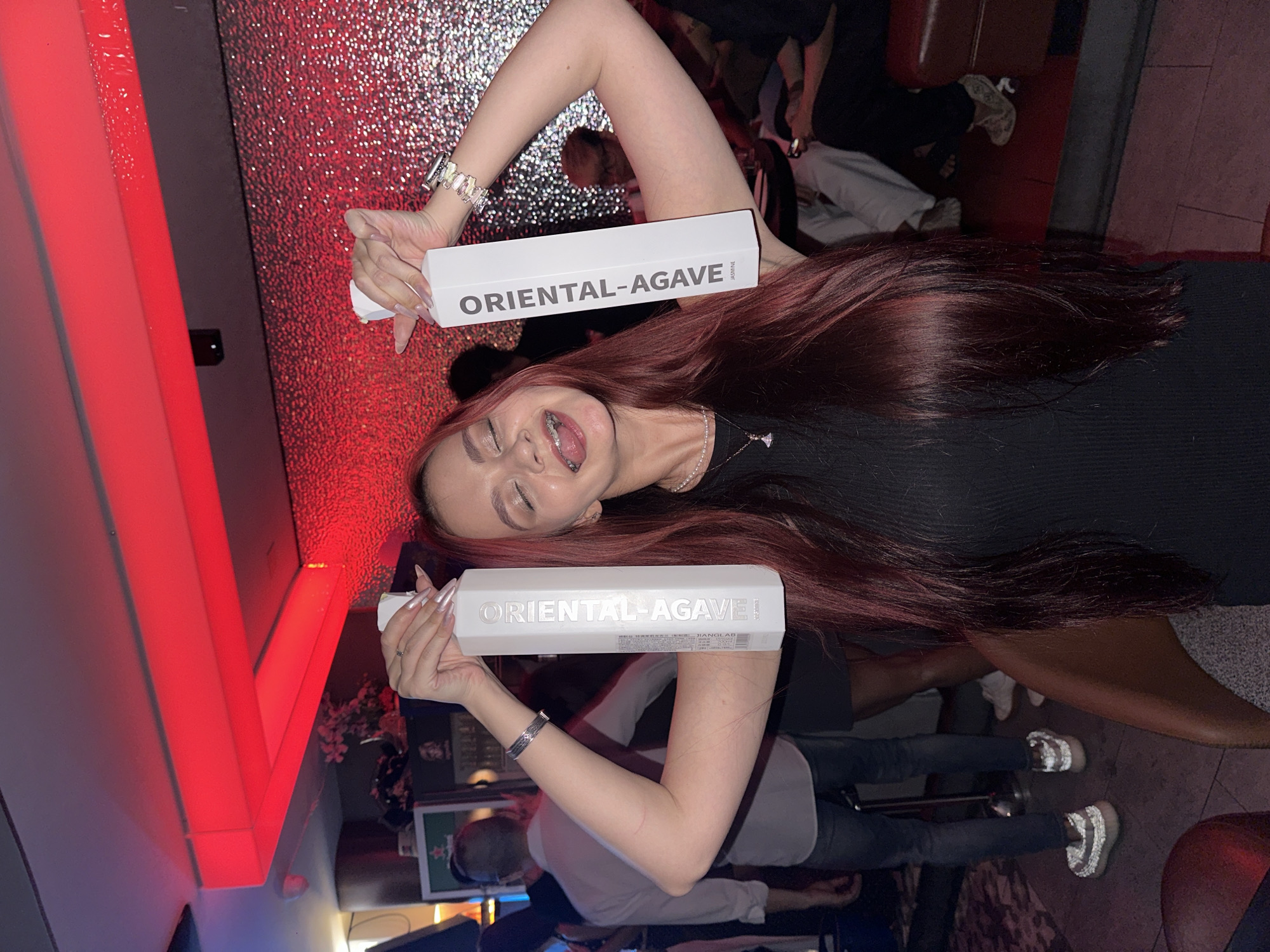 In the city of Khan Chamkar Mon, Cambodia, a young woman with long, dark red hair and braces is playfully posing for a photo in a dimly lit, possibly nightclub or bar setting. She is holding two tall, white bottles with the label "ORIENTAL-AGAVE" in large, grey lettering vertically along the side. The woman is wearing a black, sleeveless dress and several pieces of jewelry, including a bracelet on her right wrist and a necklace. She is sticking her tongue out and her eyes are closed, suggesting a lighthearted and celebratory mood. The background is blurred, but appears to be a textured, possibly red, wall. Other patrons are visible in the background, some seated and some standing, further indicating a social gathering. The lighting is predominantly red, casting a warm glow over the scene.
