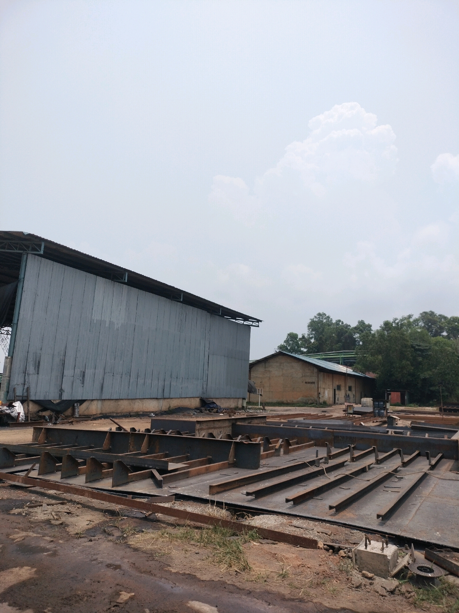 The image depicts an industrial scene in Batam, Indonesia, likely within a shipyard or construction yard. The foreground is dominated by a large, flat area covered with sections of steel, arranged in a grid-like pattern. These appear to be components for large structures, possibly ships or offshore platforms, with numerous parallel steel beams welded onto base plates.
To the left, a large, weathered corrugated metal building with a sloping roof covers a significant portion of the frame. The building's foundation is visible, and some debris, including white plastic sheeting, is present near its base.
In the mid-ground, a smaller, light brown building with a dark roof is situated amongst lush green trees. This building appears to be a more traditional structure, possibly an administrative office or a smaller workshop. Various materials and equipment, including what looks like a pallet of materials and a dark-colored truck, are visible near this building.
The sky above is overcast with large, fluffy white clouds, suggesting it is daytime with diffused sunlight. The ground is a mix of dirt, mud, and scattered debris, indicating an active and somewhat untidy industrial environment. The overall impression is one of ongoing construction or fabrication work in an outdoor setting. There are no people visible in the image.