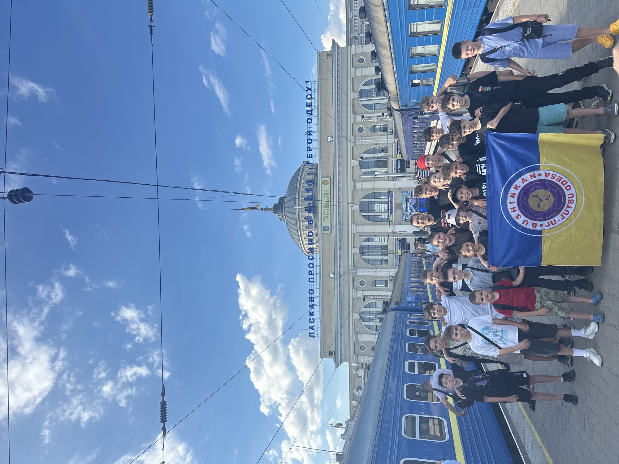 A group of young males, possibly a sports team or club, pose for a photograph on a train platform in Odesa, Ukraine. They stand in front of a bright blue train and the Odesa railway station, which features large arched windows and a prominent dome. The station building has a sign in Cyrillic script that reads "ЛАCКАВО ПРОСИМО В МІСТО ГЕРОЙ ОДЕСУ!", translating to "Welcome to the Hero City Odesa!". Several of the boys are holding up a yellow and blue flag, which appears to be a Ukrainian flag with a circular emblem that reads "BUSHINKAN ODESA". The sky is a clear blue with scattered white clouds, and overhead there are electrical wires and poles typical of a train station. The group consists of about 20 individuals, mostly teenagers, dressed in casual clothing. Some are smiling and looking at the camera, while others have their arms around each other. The overall scene suggests a departure or arrival at the train station, with a sense of camaraderie and pride among the group.