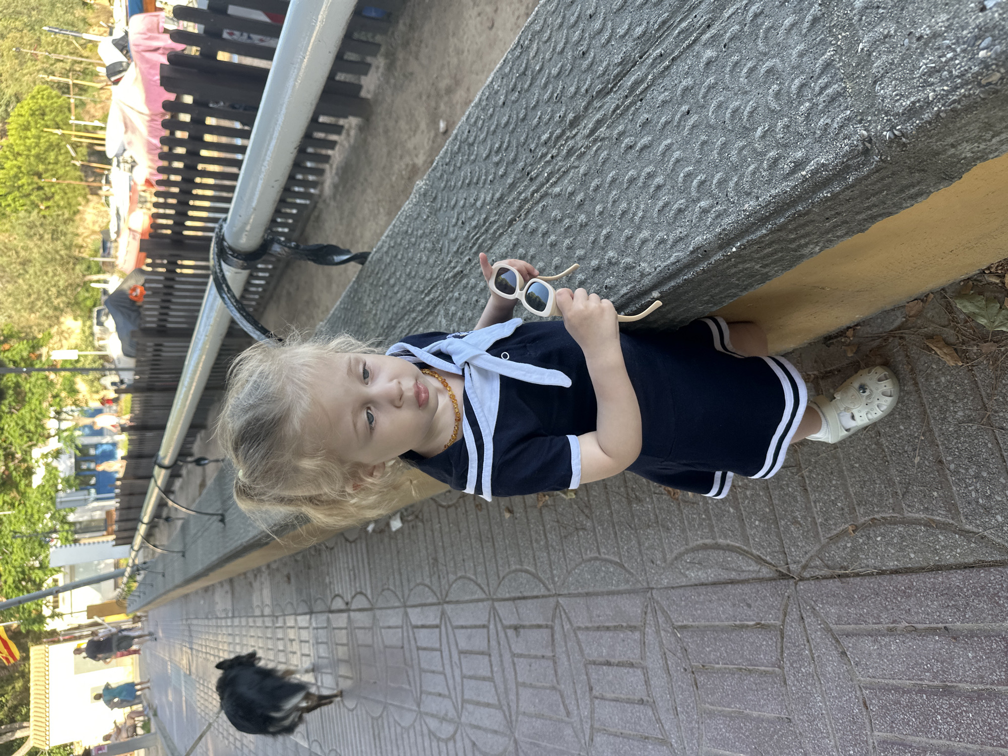 The image shows a young girl standing on a paved walkway, holding a pair of sunglasses. She is wearing a navy blue dress with white trim and a white sailor collar, and a small amber necklace. Her hair is blonde and she is looking towards the camera with a neutral expression. Behind her, a black dog is walking away from the camera. The walkway is bordered by a textured concrete wall and a yellow painted curb. In the background, there is a black metal fence and lush green trees. Further beyond the fence, there are hints of buildings and other people, suggesting a public space like a promenade or park. The lighting suggests it is daytime, possibly late afternoon due to the warm, soft light. The setting appears to be outdoors in a sunny climate, consistent with a Spanish coastal town like Sant Feliu de Guíxols. There is no visible text in the image.