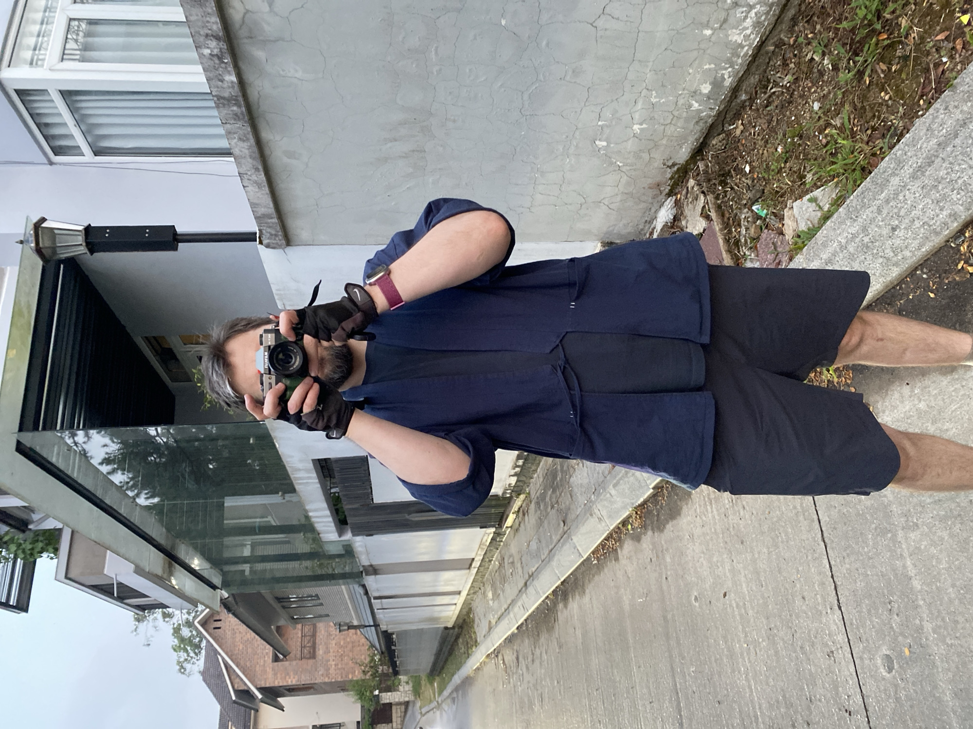 A man stands on a concrete sidewalk facing a camera, holding a black camera at eye level. He wears a navy blue shirt, black shorts, and black gloves, with a red watch on his left wrist. His gray hair is visible, and his beard is short. The backdrop includes a street leading to houses of varying architectural styles, some with glass facades and others with brick or light-colored exteriors. To the left is a multi-story building with a light gray exterior and a window with blinds. The sidewalk borders a thin strip of soil with greenery and a concrete barrier. The scene is located in Da Lat, Vietnam.