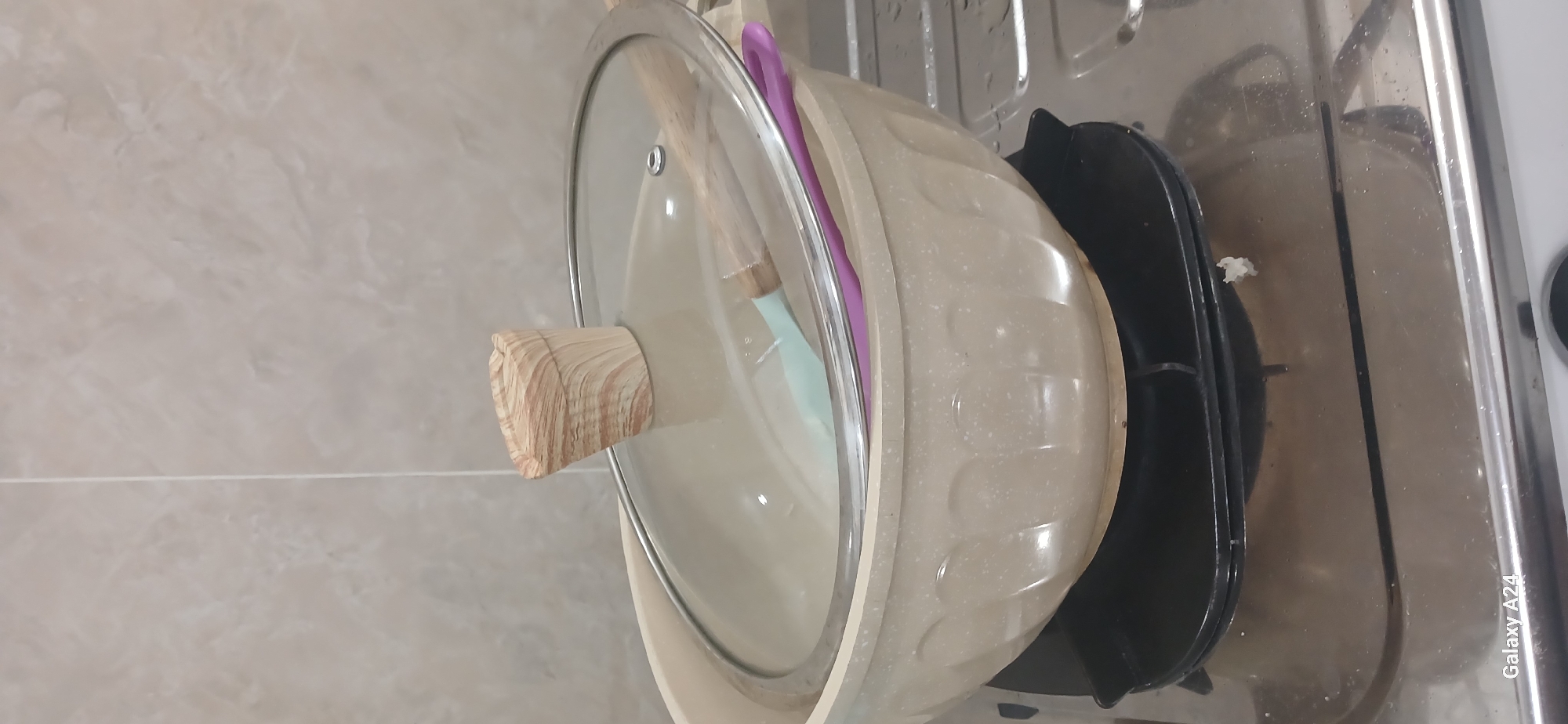 A close-up, eye-level shot shows a beige pot with a glass lid on a gas stove. The pot has decorative fluting around its sides. Inside the pot, visible through the glass lid, are two kitchen utensils: a light green spatula and a purple spatula. The lid's handle is a light brown, wood-grain pattern. The stove's burner grate is black, and the stovetop is stainless steel with some water droplets and a small piece of white debris on it. The background is a tiled wall with a light beige, textured pattern. The image is taken indoors, likely in a kitchen setting. The overall scene appears to be in Kuwarisan, Indonesia.