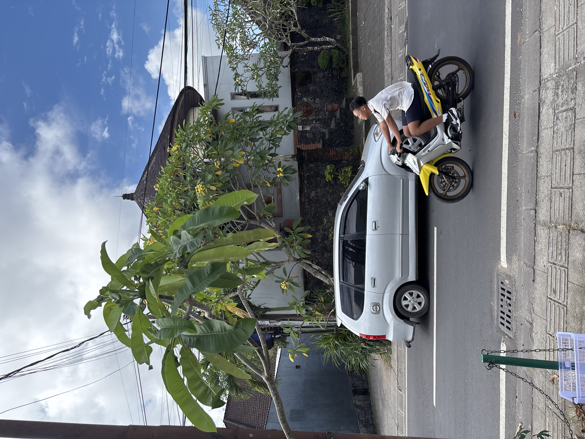 A street scene in Kuta Selatan, Indonesia, shows a white car parked along the roadside. A person in a white shirt and dark shorts is on a yellow and white motorcycle parked next to the car, appearing to interact with it. The street is paved with concrete and has a white line marking a parking space. A tree with large green leaves is visible in front of a building with a dark roof and white walls. Overhead wires are visible against the blue and cloudy sky. A curb, a storm drain, and part of a basket attached by chains to a green pole are visible at the edge of the street.