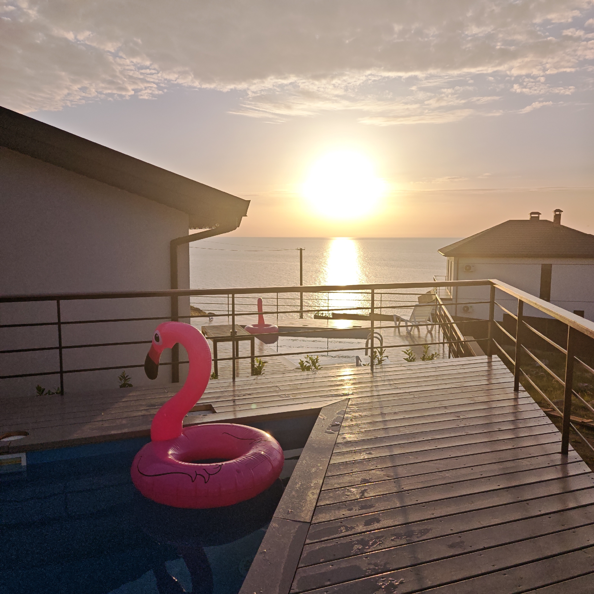 A poolside scene at sunset features a pink flamingo inflatable floating in a pool. The pool is bordered by a wooden deck with metal railings. In the background, the sun is setting over the ocean, visible between two residential buildings. A table with a second flamingo float is placed by the railing.