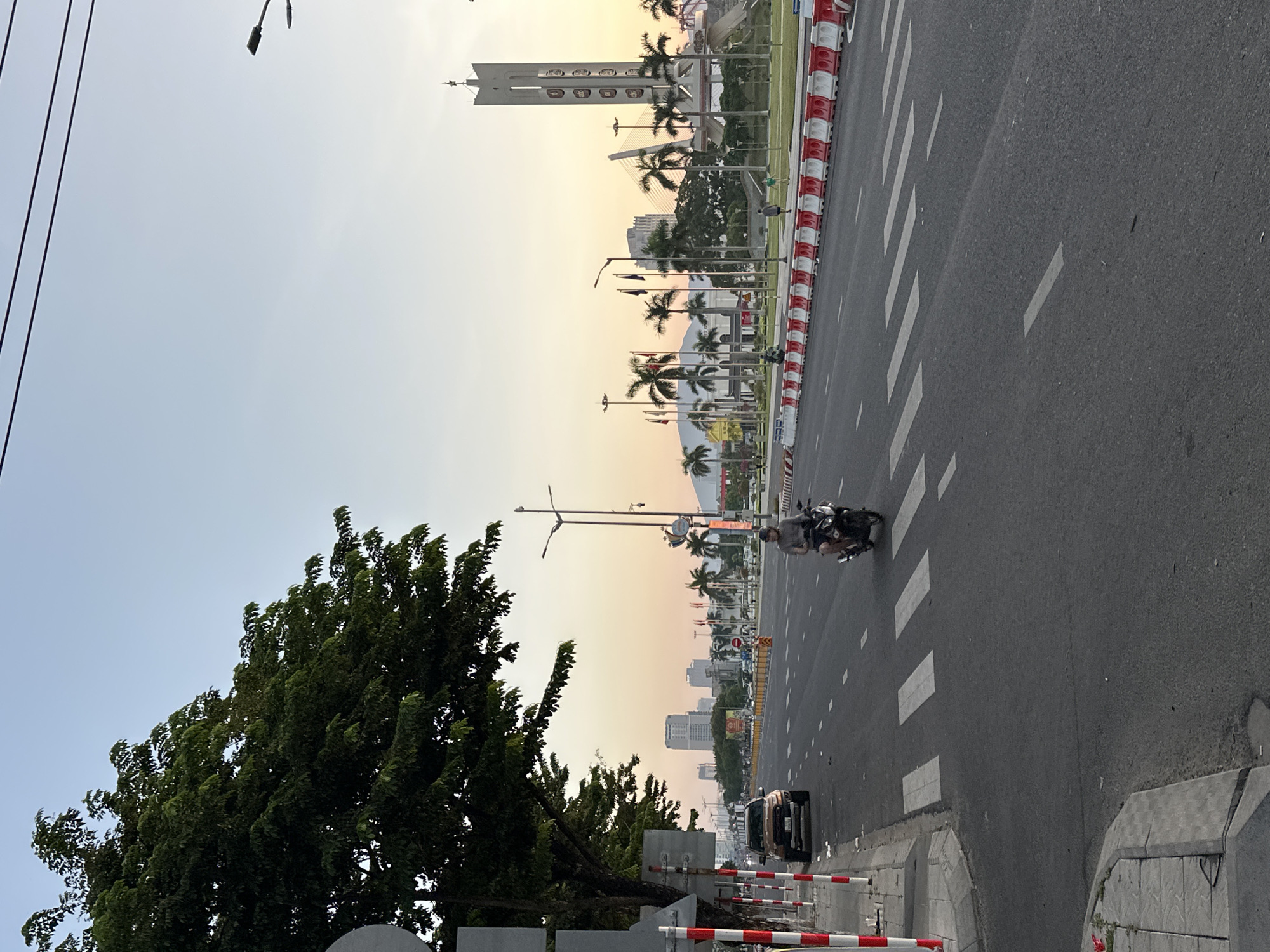 A street scene in Da Nang, Vietnam, likely near Dragon Bridge and the Han River, is captured at dusk. The sky displays a gradient of pale orange to light blue, indicating sunset. A wide, multi-lane road dominates the foreground, with white dashed lines marking the lanes and a prominent crosswalk. A lone motorcyclist, wearing a helmet, is in motion on the right side of the road, heading towards the viewer. A silver SUV is parked on the left side, near a sidewalk lined with red and white bollards.
To the right of the road, a landscaped area features palm trees and several flags. A tall, modern, tower-like structure with decorative panels stands prominently, likely a monument or landmark. In the distance, several tall buildings rise against the horizon, suggesting an urban environment. A cable-stayed bridge is visible in the background, partially obscured by trees. The overall atmosphere is calm, with a few pedestrians walking in the landscaped area. The lighting suggests the transition from day to evening.