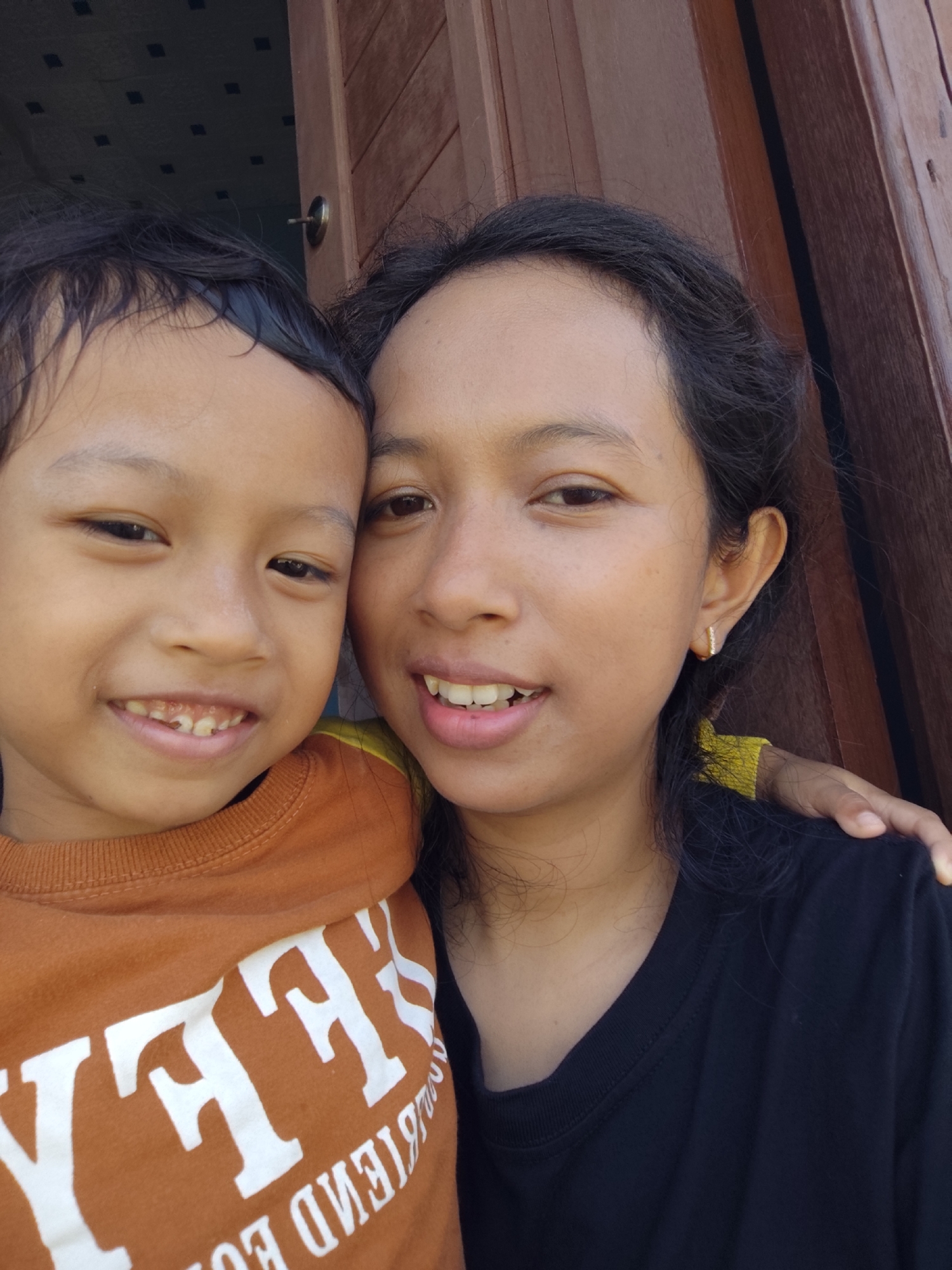 A head and shoulders shot shows two people posing for a selfie. A young boy on the left, facing the camera, smiles broadly, revealing his teeth. He wears an orange t-shirt with white lettering. Beside him, on the right, a young woman is also facing the camera, smiling. She has a small earring in her right ear and wears a black t-shirt. Her left arm is placed on the boy's shoulder. The background appears to be a wooden doorframe.