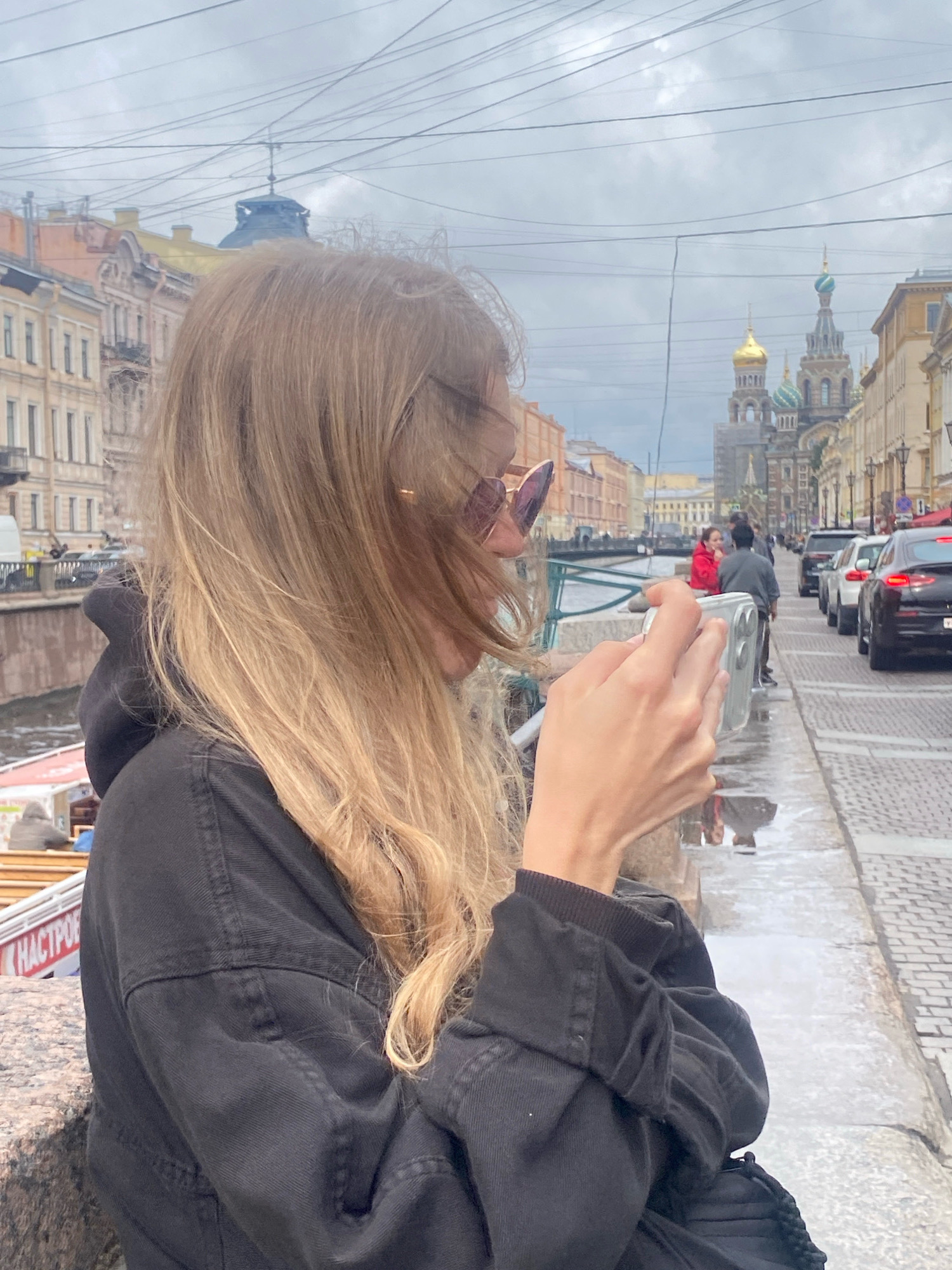 A young woman with blonde hair and sunglasses is holding a silver smartphone and taking a picture in a city. She is wearing a black jacket and standing on a stone embankment next to a canal. In the background, there are buildings with a golden dome and a church with colorful domes, characteristic of Saint Petersburg, Russia. Cars are parked along the street next to the canal, and there are overhead wires visible against the cloudy sky. The canal water is brown, reflecting the surrounding architecture and the sky. There are a few other people in the background, some walking and some near a boat.