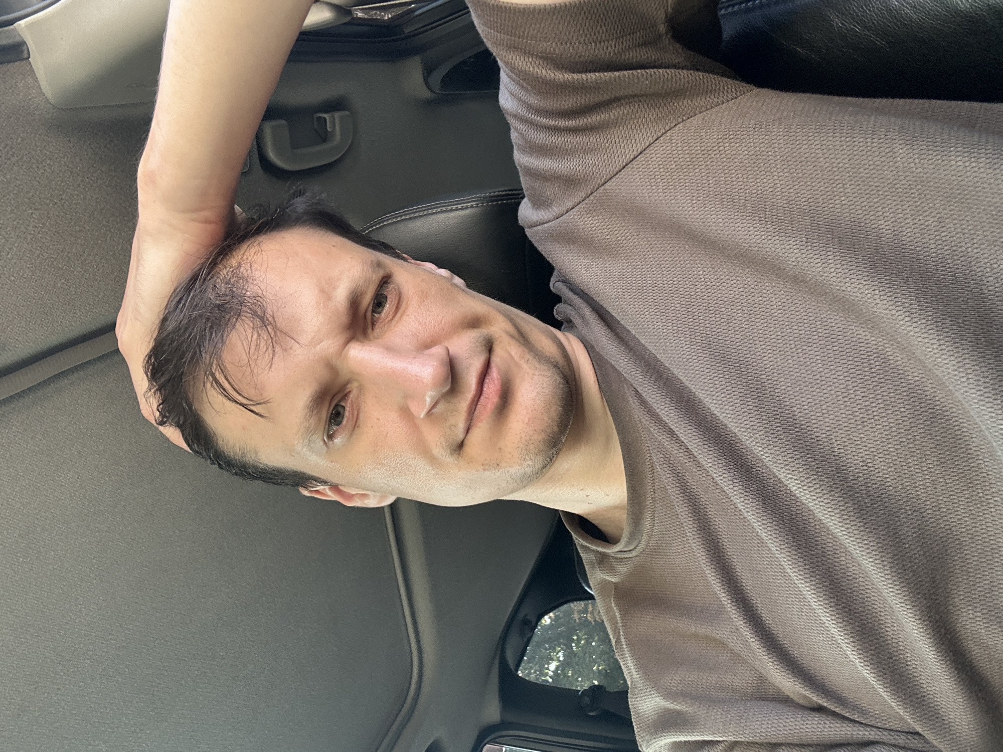 A man is sitting in the driver's seat of a car, with his right arm raised and hand resting on the back of his head. He is looking directly at the camera. The car appears to be parked, as suggested by the static posture and the man's relaxed demeanor. The interior of the car is visible, including the dark leather seat and the car's headliner, which has a sunroof. The man is wearing a brown, textured, long-sleeved shirt. The background outside the car shows foliage, indicating a green, possibly park-like or tree-lined area. The lighting suggests it is daytime. There is no information or visual evidence to suggest the location is the city of Kamianets-Podilskyi, Ukraine.