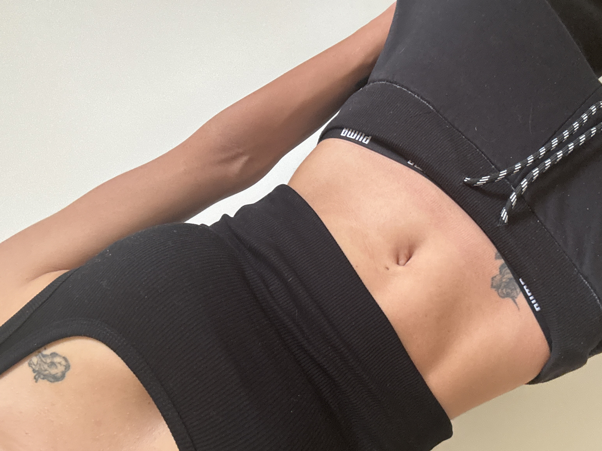 The image presents a close-up, angled view of a person's torso, arm, and midsection, set against a plain, light-colored background. The person is wearing black athletic or loungewear. The upper garment is a black, ribbed crop top or sports bra, and the lower garment appears to be black shorts with a ribbed waistband and black and white striped drawstrings.
Visible text on the elastic bands of both the top and the shorts identifies the brand as "PUMA," though it appears inverted in the image as "AMUP." The person's midriff is exposed, revealing a navel. Two tattoos are present: a small, detailed tattoo, possibly a cherub or similar figure, is located on the left side of the torso near the rib cage. Another tattoo is partially visible on the right hip area, just above the waistband of the shorts.
The skin tone is warm, and the lighting is soft and diffused. The pose suggests a relaxed or styled shot, possibly a selfie, focusing on the clothing and body art. The plain background implies an indoor setting, likely a room or studio. No specific activity beyond posing for the photograph is depicted.
