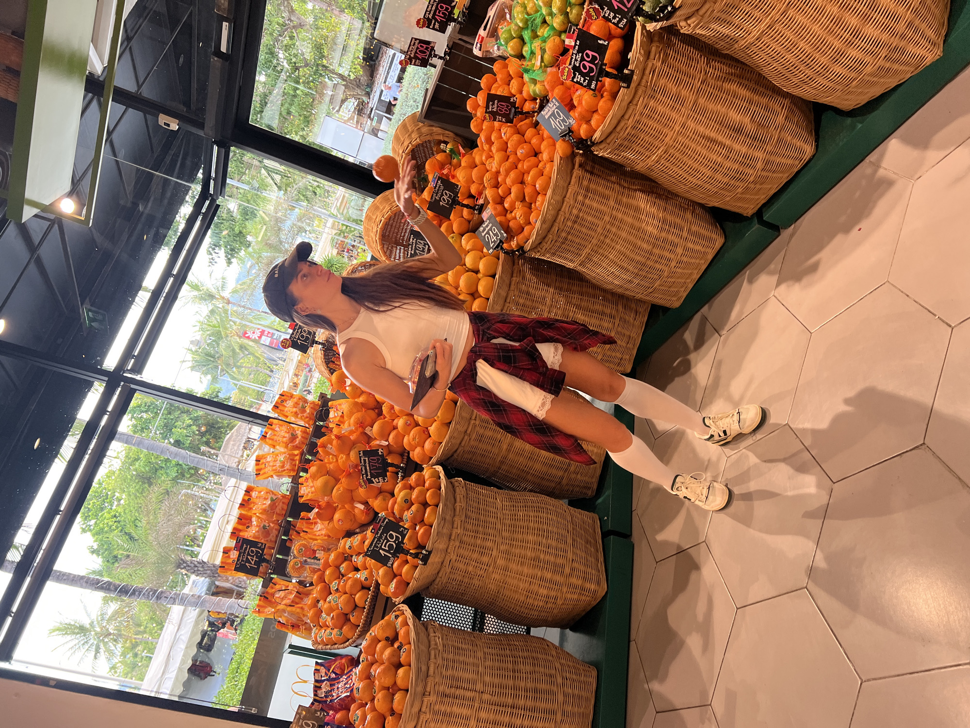 Here is a description of the image:
The image shows a woman standing in a store with large baskets filled with oranges. The store appears to be located in Baan Chaweng, Thailand, based on the visible surroundings outside the large glass windows. The woman wears a white tank top, a red plaid shirt tied around her waist, short white socks, and sneakers. She's holding an orange in her hand, seemingly juggling it, and she is also holding a phone or small device in the other hand. Baskets of oranges with price tags are arranged behind her on green shelves. Additional baskets of fruit can be seen in the background, along with the exterior view of palm trees and what looks like a building or structure near a beach. The floor is tiled with large gray hexagonal tiles.