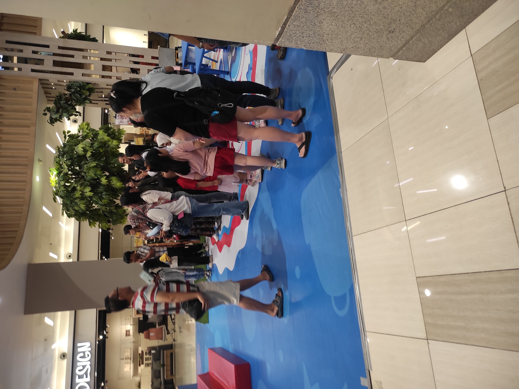 A group of people are gathered on a blue mat in what appears to be an indoor shopping mall in Surabaya, Indonesia. The mat has a colorful, wavy design. To the left, there is a red tiered structure. In the background, a large, leafy tree is visible, and a sign that reads "DESIGN" is mounted on a wall. Several individuals, including adults and children, are standing and looking in various directions. One man in a striped shirt is walking away from the camera. The floor surrounding the blue mat is tiled.