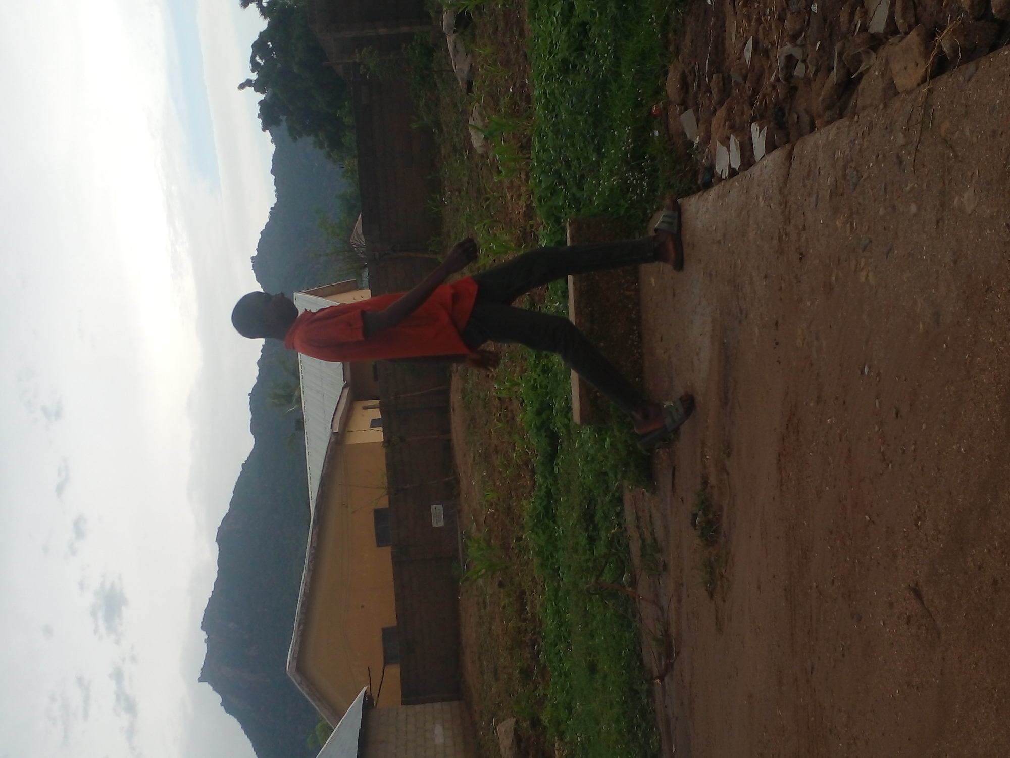 A young Black male, wearing a red t-shirt and dark trousers with sandals, walks to the right on a paved surface. He is in profile, facing right, with his head turned to look forward. In the background, there is a yellow building with a metal roof, a brick wall, and a patch of cultivated land with young plants. Beyond the buildings, a large, rocky mountain range is visible against a cloudy sky. The foreground is a dirt path. The scene appears to be in a rural or semi-rural area of Langtang, Nigeria.