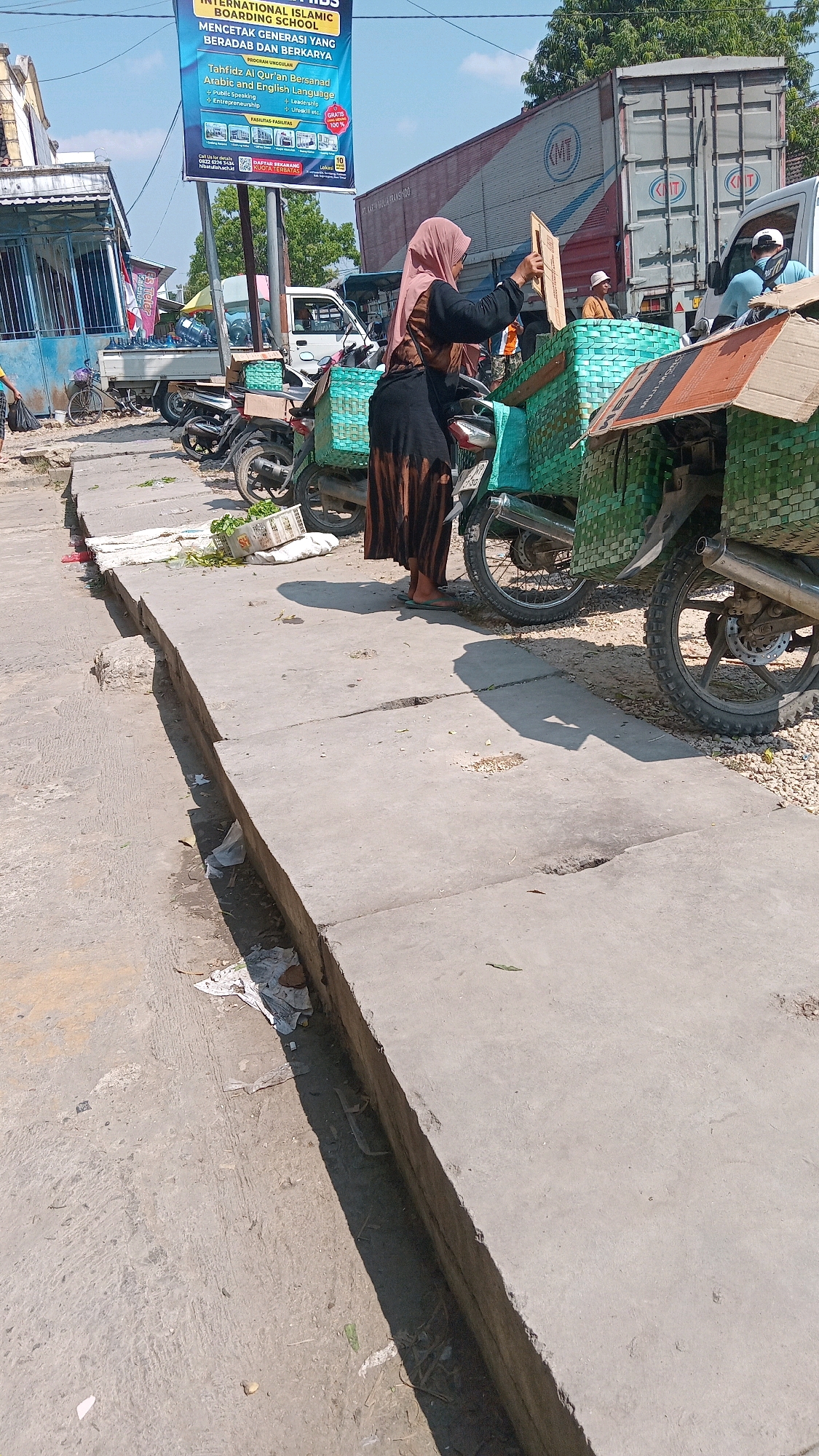 In Kunduran, Indonesia, a woman wearing a pink hijab and a long brown and black patterned skirt is holding a flat, rectangular wooden object while standing next to a motorcycle with a green woven basket attached. The motorcycle is parked on a concrete surface, next to a larger concrete drainage ditch filled with debris. In the background, other motorcycles and vehicles are visible, along with a large shipping container truck and a white pickup truck. A blue banner advertising an Islamic school is also prominent in the background. The scene appears to be an outdoor market or roadside area, with a bright, sunny sky.