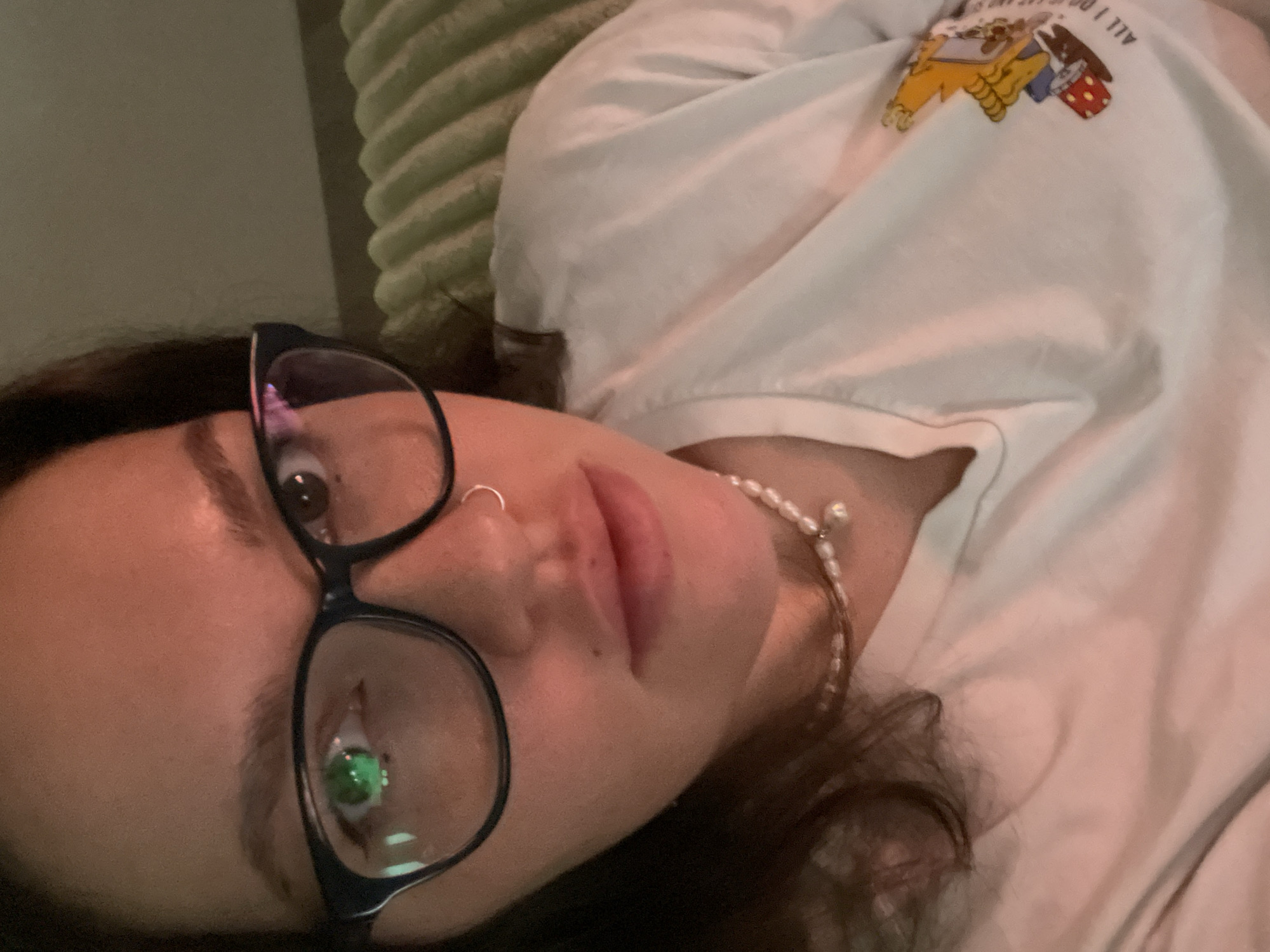 An indoor, close-up shot captured in Izhevsk, Russia, depicts a young individual, likely female, lying down and looking directly at the camera. The person has dark hair, wears black-framed glasses, and a silver hoop nose ring on their left nostril. A delicate pearl necklace or choker is visible around their neck.
They are wearing a white t-shirt featuring a colorful graphic design of cartoon characters, with the partially visible text "ALL I DO IS..." and "...S" above it. The person's expression appears neutral or contemplative, with slightly parted lips. The lighting is dim and soft, suggesting an evening or nighttime setting. Behind the person, a light green, ribbed or textured pillow or blanket is visible, indicating they are likely on a bed or sofa. Reflections can be seen in the glasses, with a distinct green light reflected in the lower left lens. The image is framed from a slightly tilted, eye-level perspective.