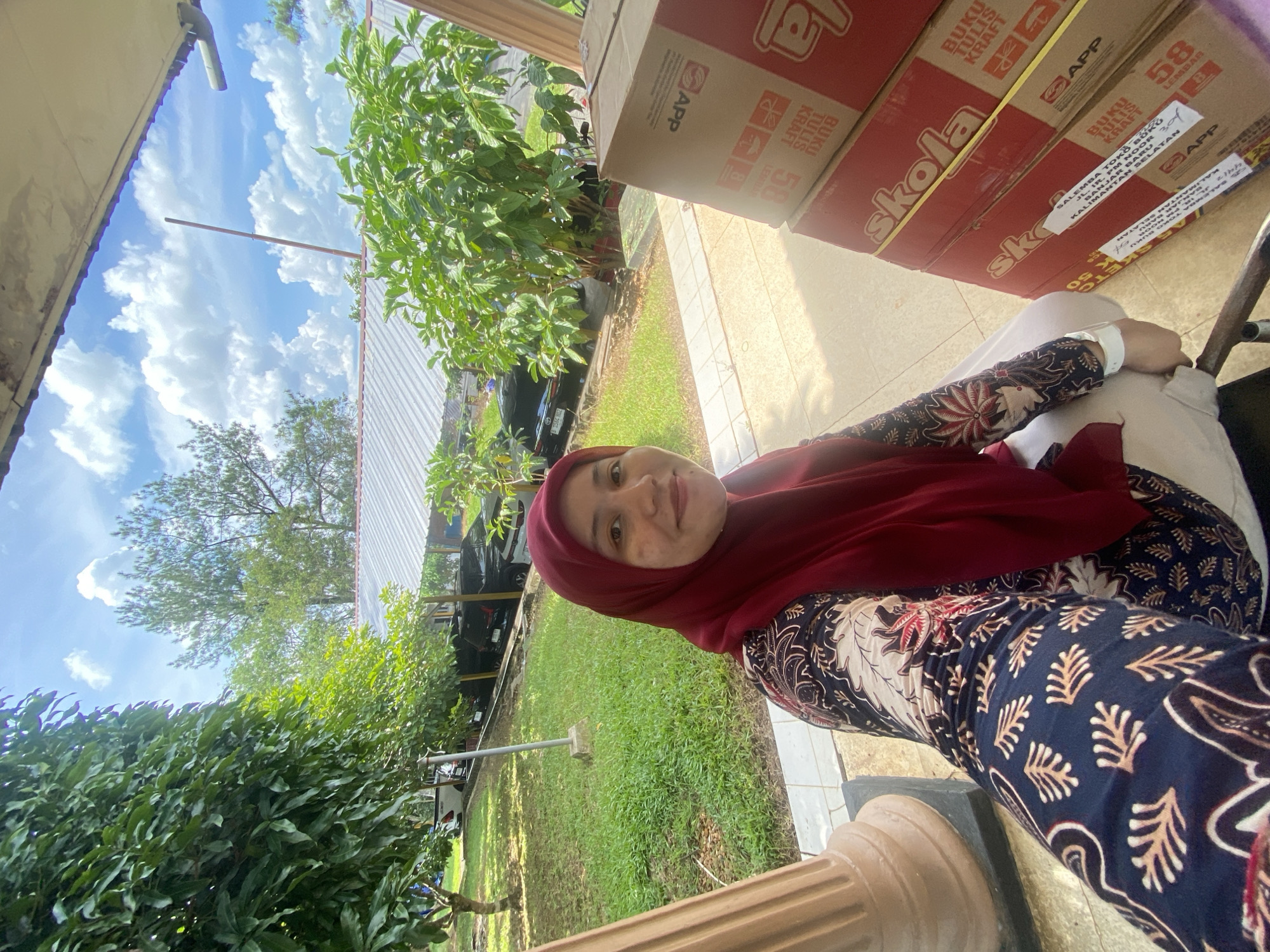 In Banjarbaru, Indonesia, a woman wearing a maroon hijab and a batik patterned top is captured in a selfie. She is seated next to a stack of red boxes, some of which have "Skola" and "APP" branding. The scene appears to be outdoors, possibly on a porch or covered area, with lush green grass, trees, and a cloudy blue sky visible in the background. Several cars are parked in the distance under a covered structure.
