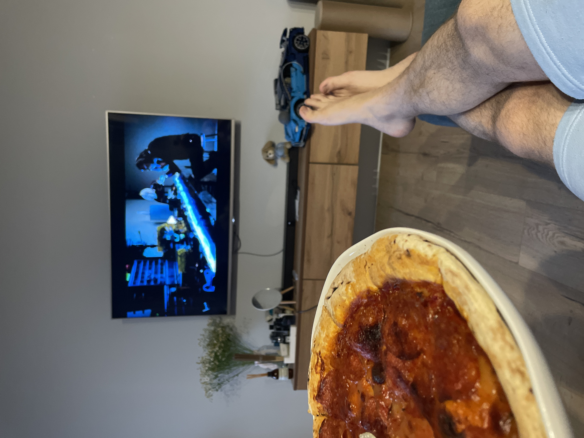 The image shows a person reclining with legs crossed, watching TV and with a pizza in the foreground. The TV displays a scene from the movie *Star Wars*. A blue toy car and a small plush animal are on the TV stand. A vase with baby's breath is visible on the stand below. The location is likely indoors. No specific indication suggests Krasnoyarsk, Russia.